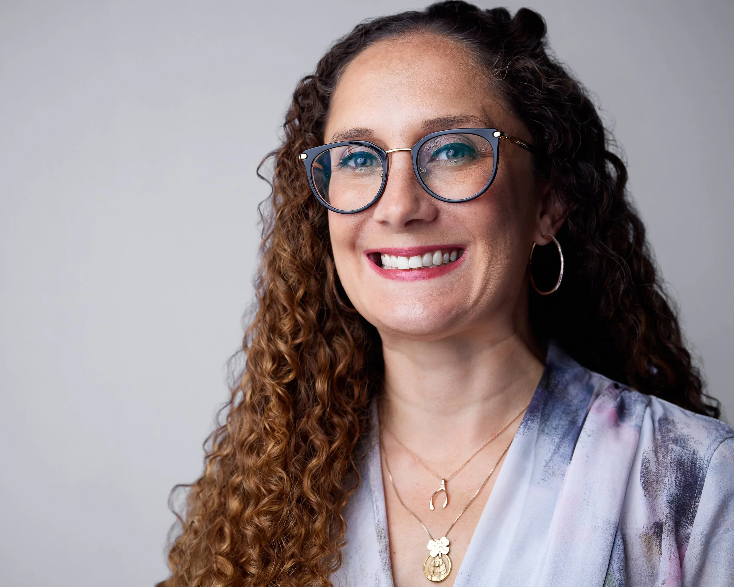 Close-up of a woman with long curly hair, wearing glasses, earrings, and layered necklaces, smiling against a plain light gray background.