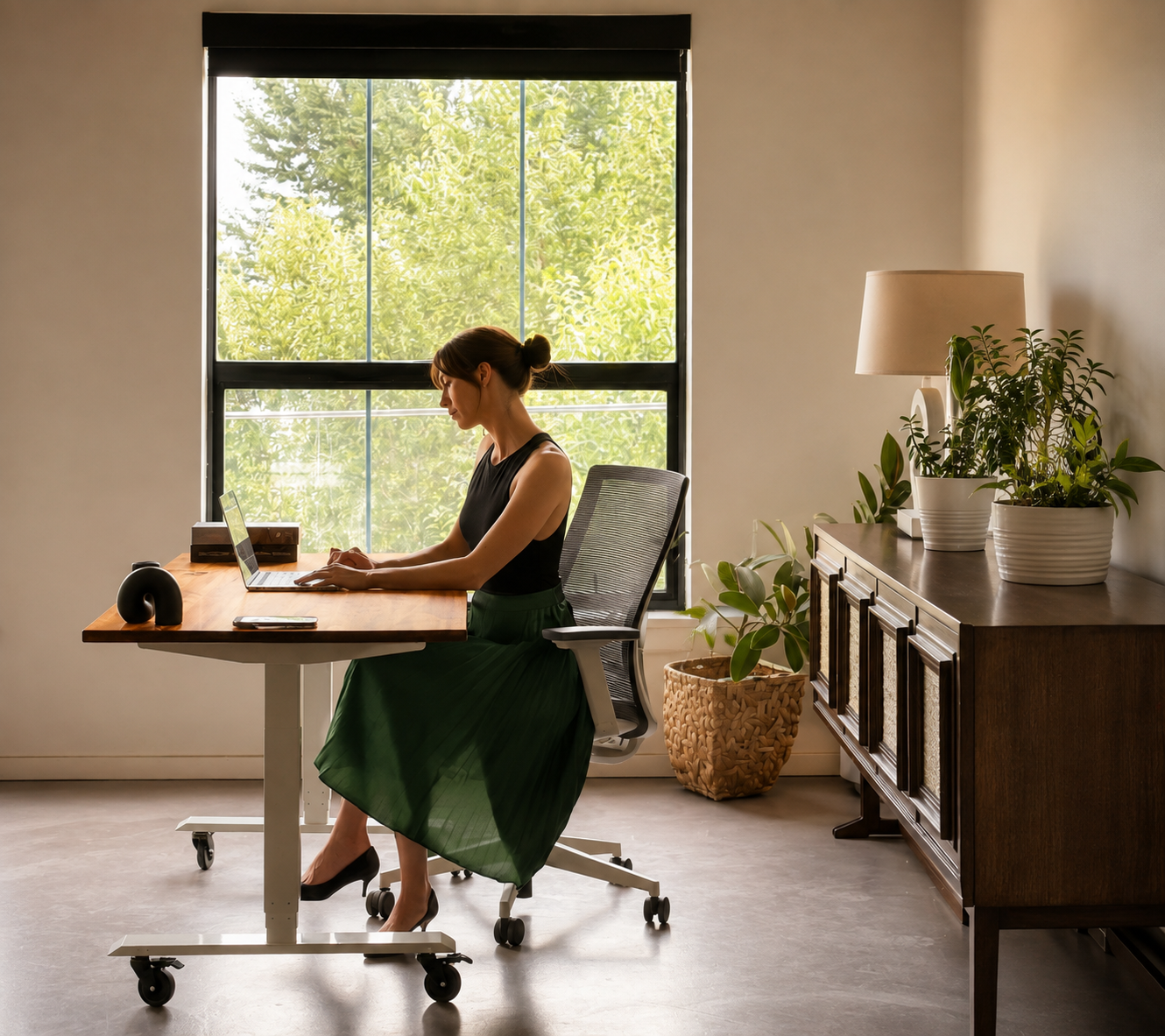 A woman working on a laptop at a desk in a bright room with large window, plants on sideboard, and neutral decor.