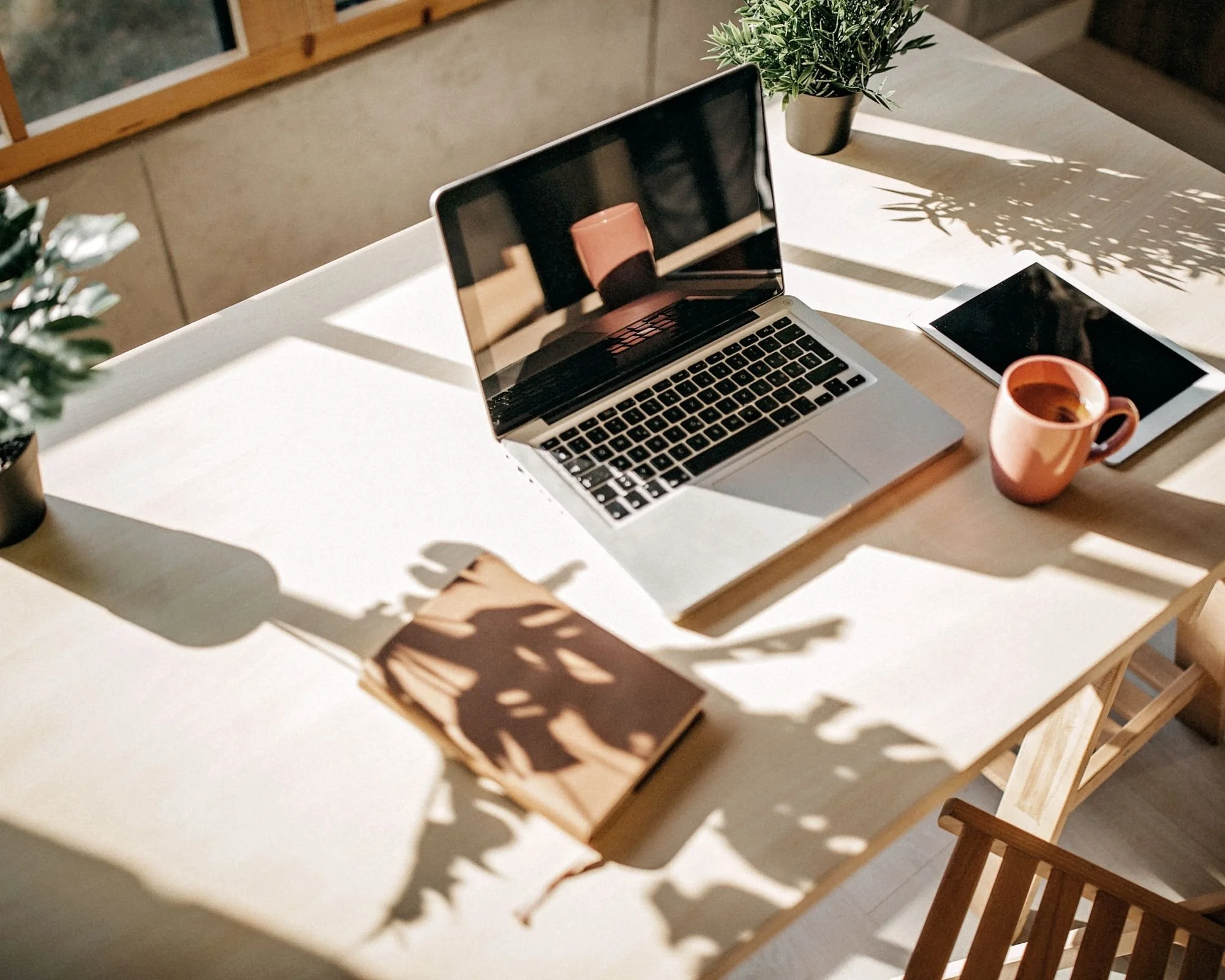 A white table with a laptop, a tablet, two potted plants, a coffee mug, and a stack of brown paper envelopes, with sunlight casting shadows.