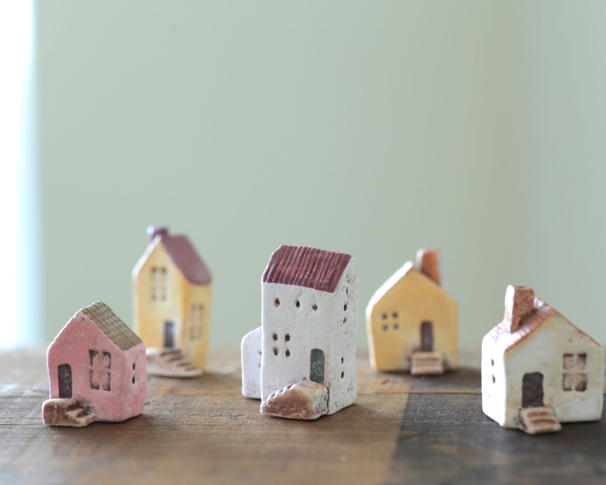 Several small ceramic house figurines with various colors and designs, arranged on a wooden surface against a muted green background.