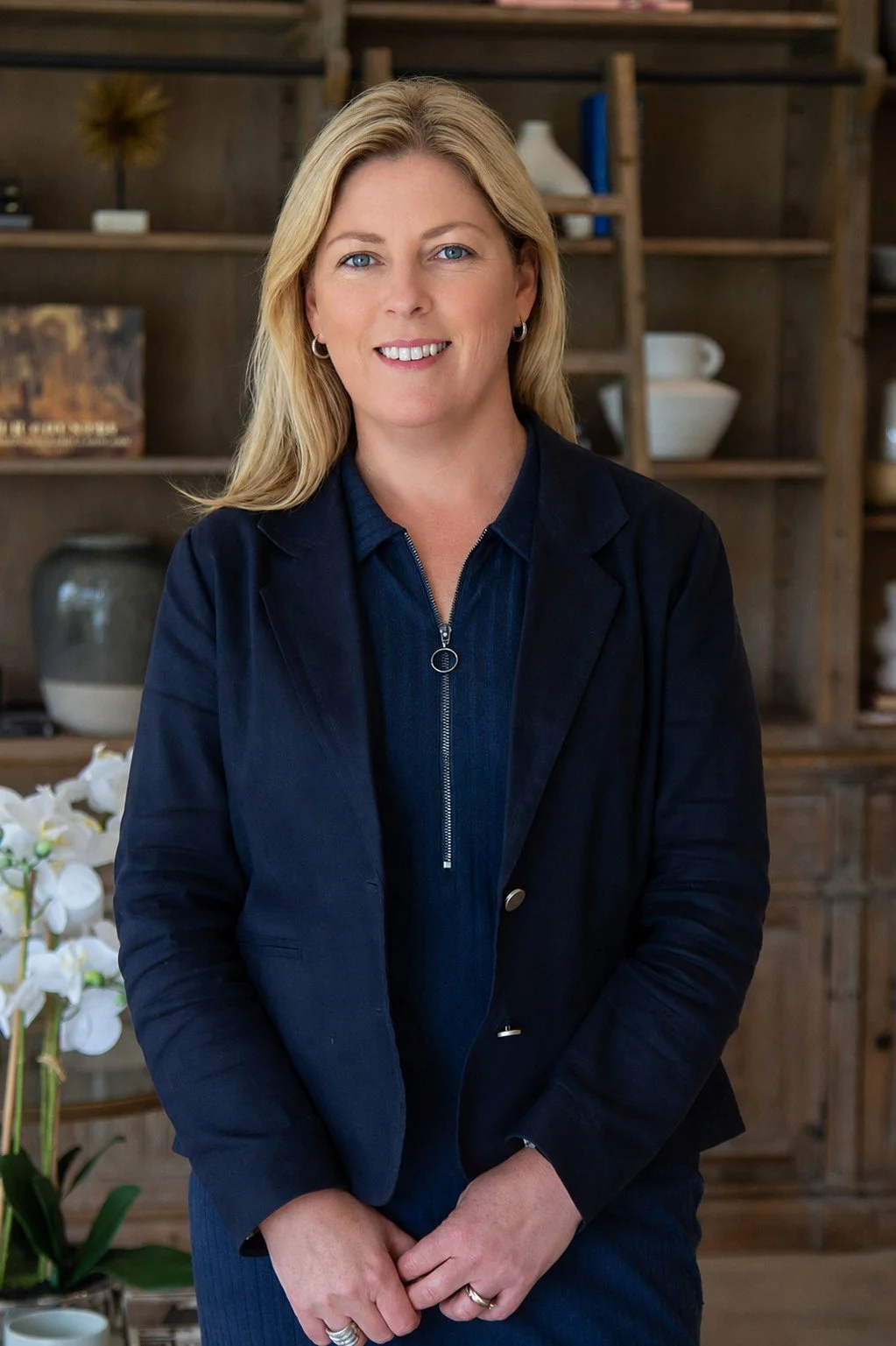 A woman with blonde hair and blue eyes smiling, dressed in a dark blue blazer and zip-up shirt, standing indoors with a wooden shelf filled with decorative items and white flowers in the background.