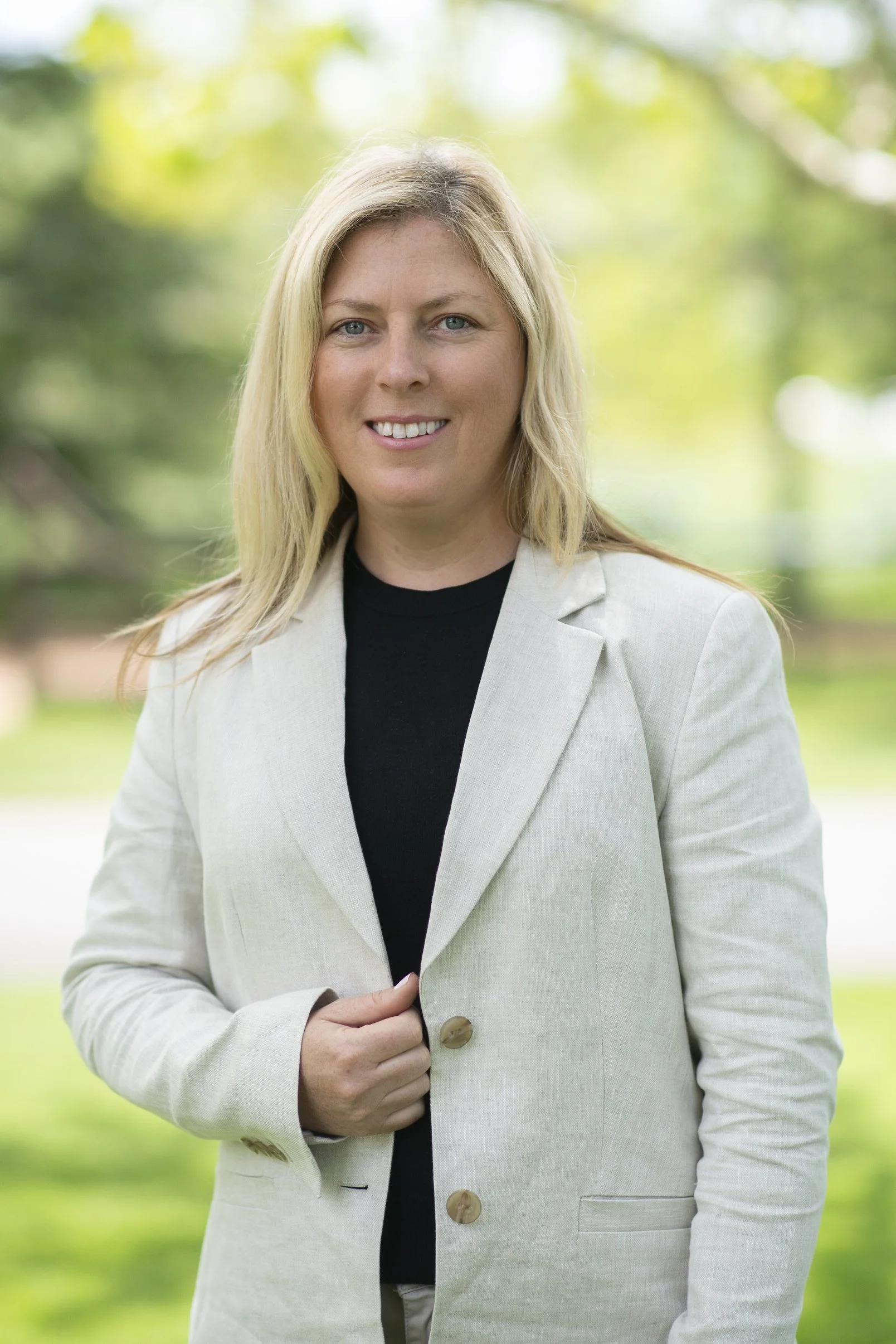 A blonde woman in a light-colored blazer and black shirt standing outdoors with a blurred green background.