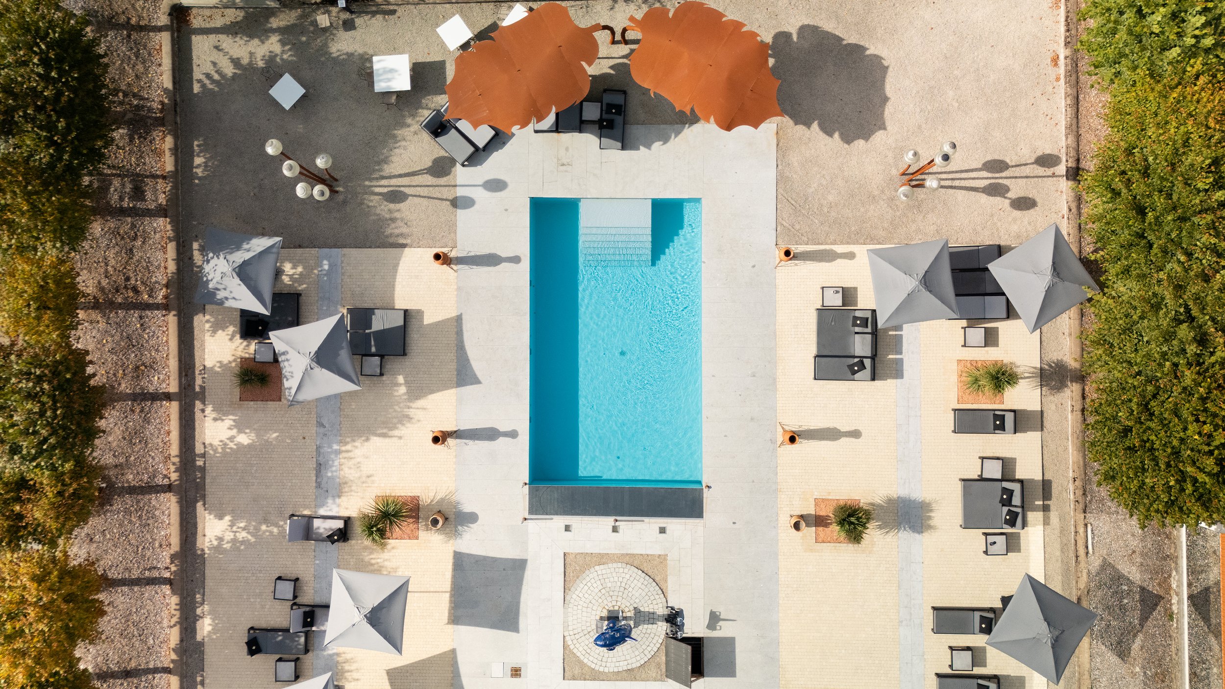 Vue aérienne d'une piscine rectangulaire dans une zone de détente avec parasols, chaises longues, et arbres entourant la zone.