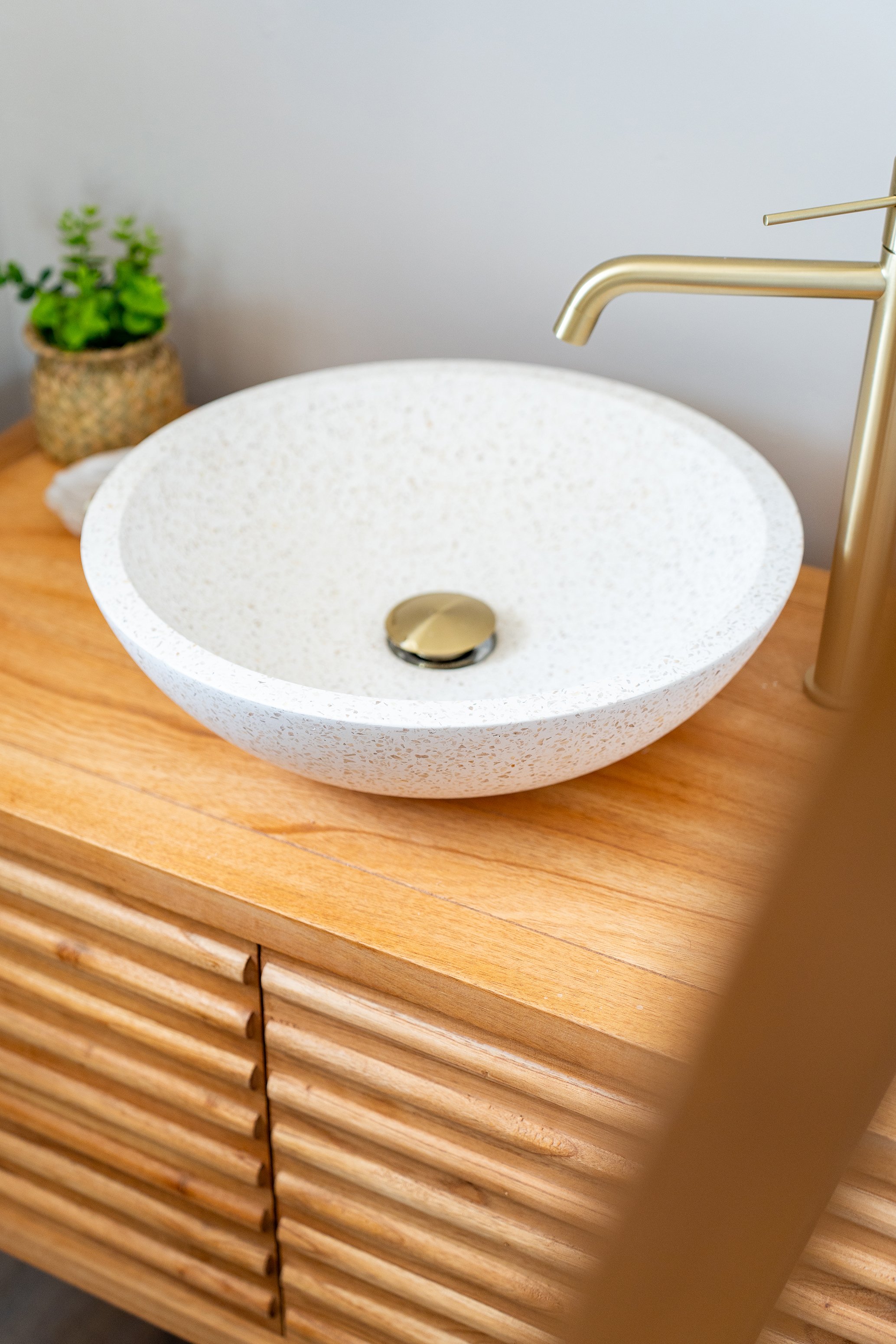 Un lavabo en pierre ou composite blanc avec un robinet en métal doré, placé sur une console en bois clair, avec une petite plante verte en pot en ardoise à gauche.