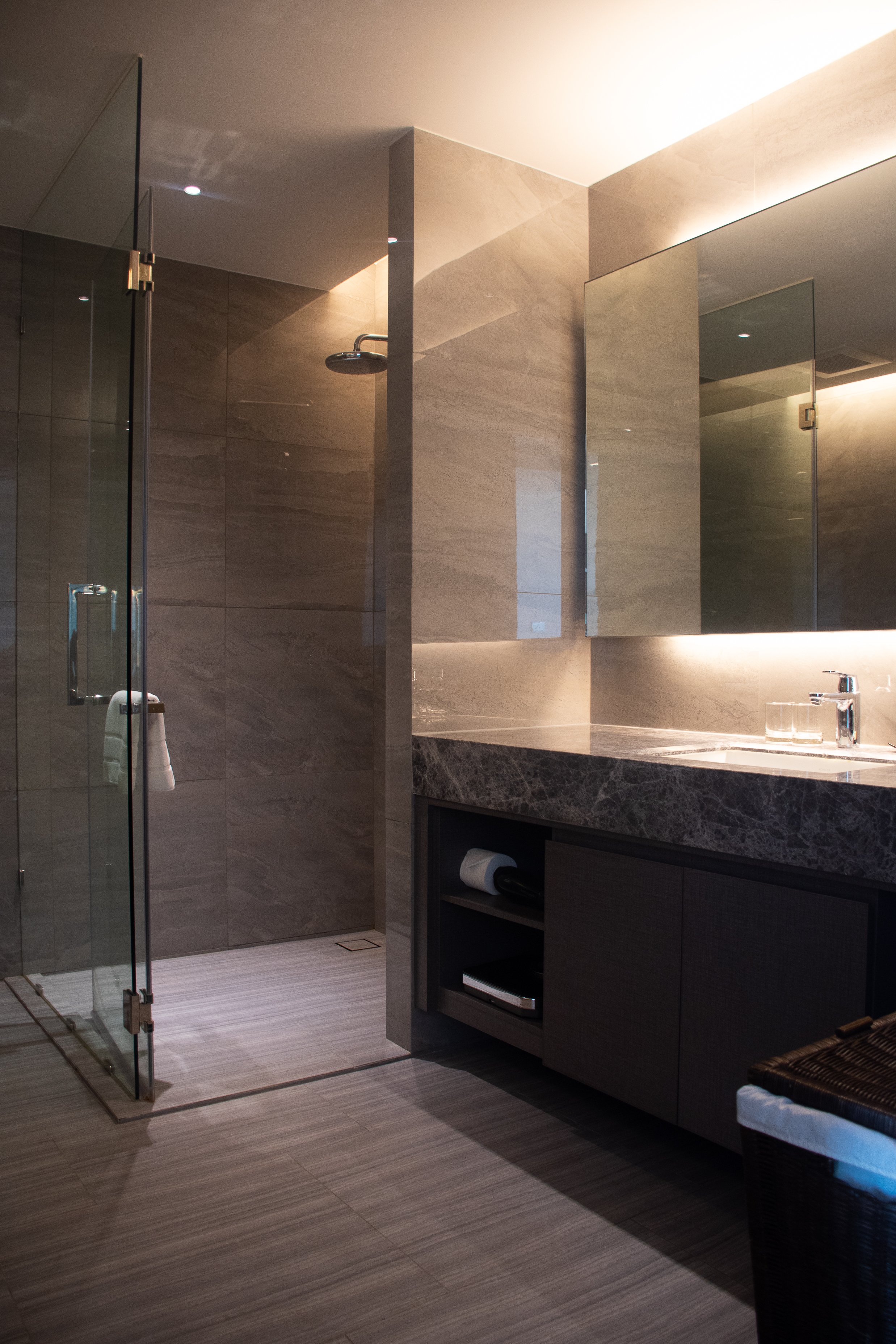 Une salle de bain moderne avec une douche en verre, un lavabo avec un miroir, une armoire noire et un panier en rotin, décorée avec des murs en pierre beige et un éclairage tamisé.