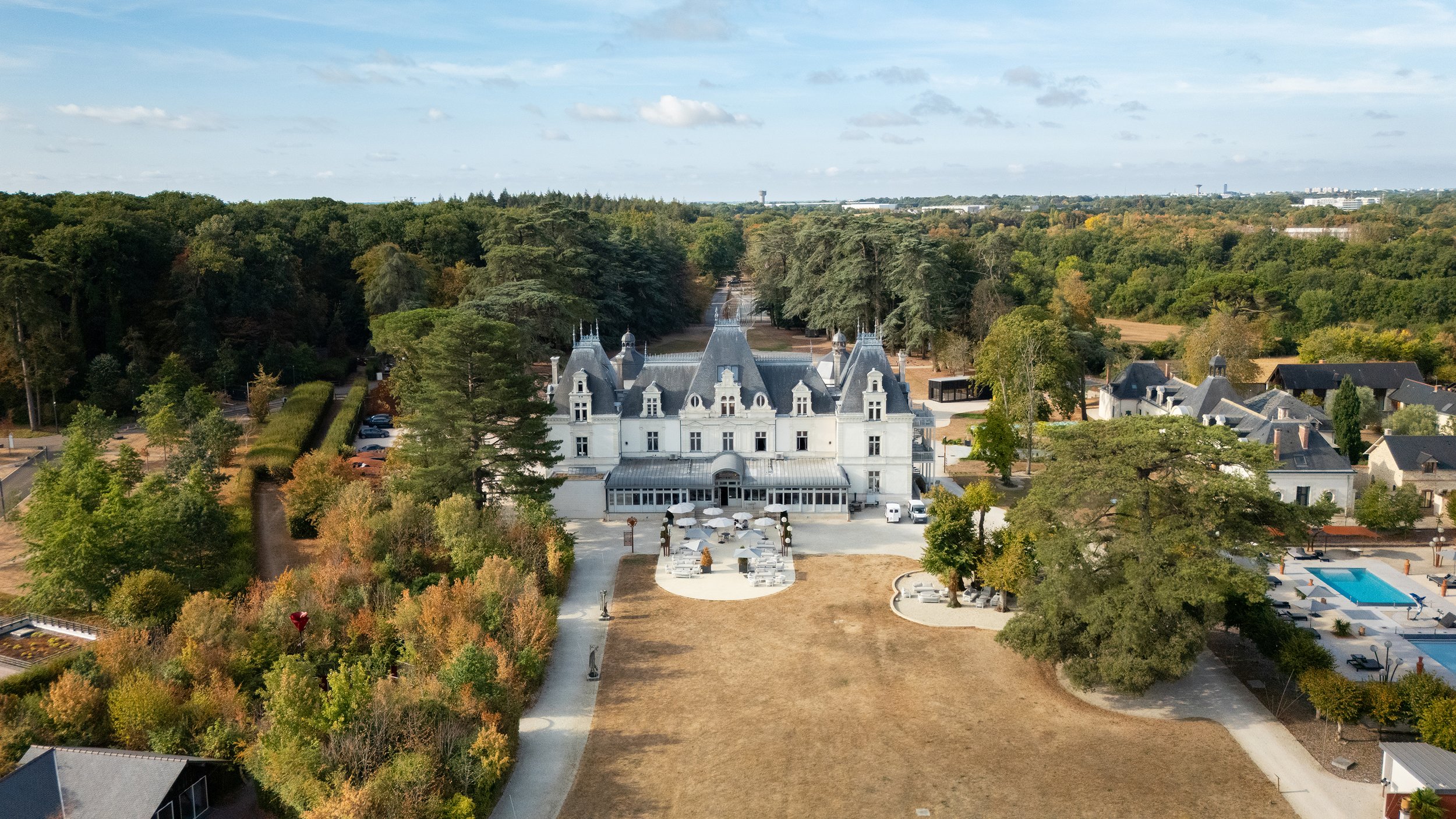 Vue aérienne d'un grand château blanc entouré d'arbres, avec une terrasse extérieure équipée de parasols et un bâtiment piscine à proximité, dans un environnement verdoyant et urbain.