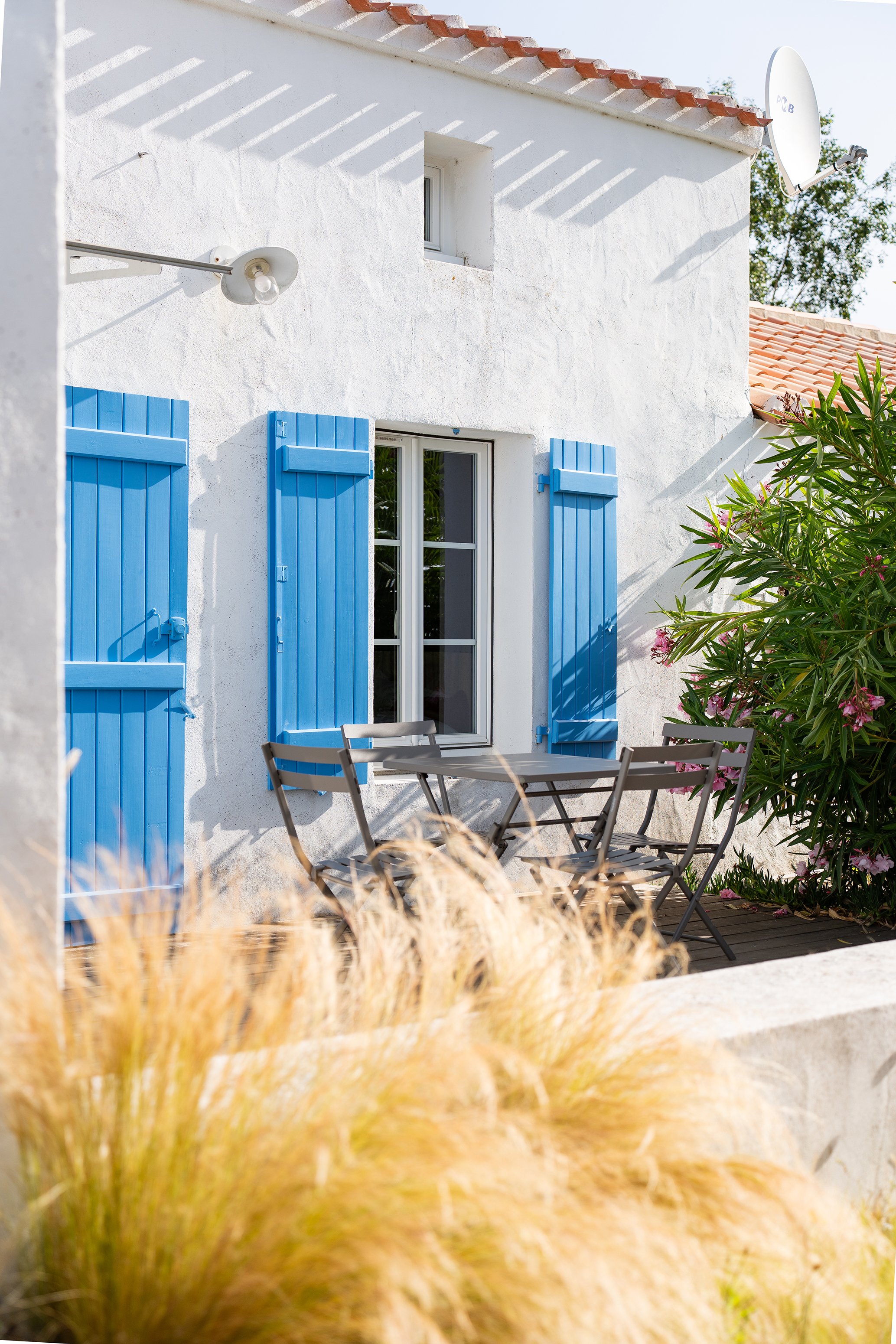 Façade blanche d'une maison avec volets bleus, un panneau solaire et un éclairage extérieur, vue partielle depuis un jardin avec des plantes ornementales.