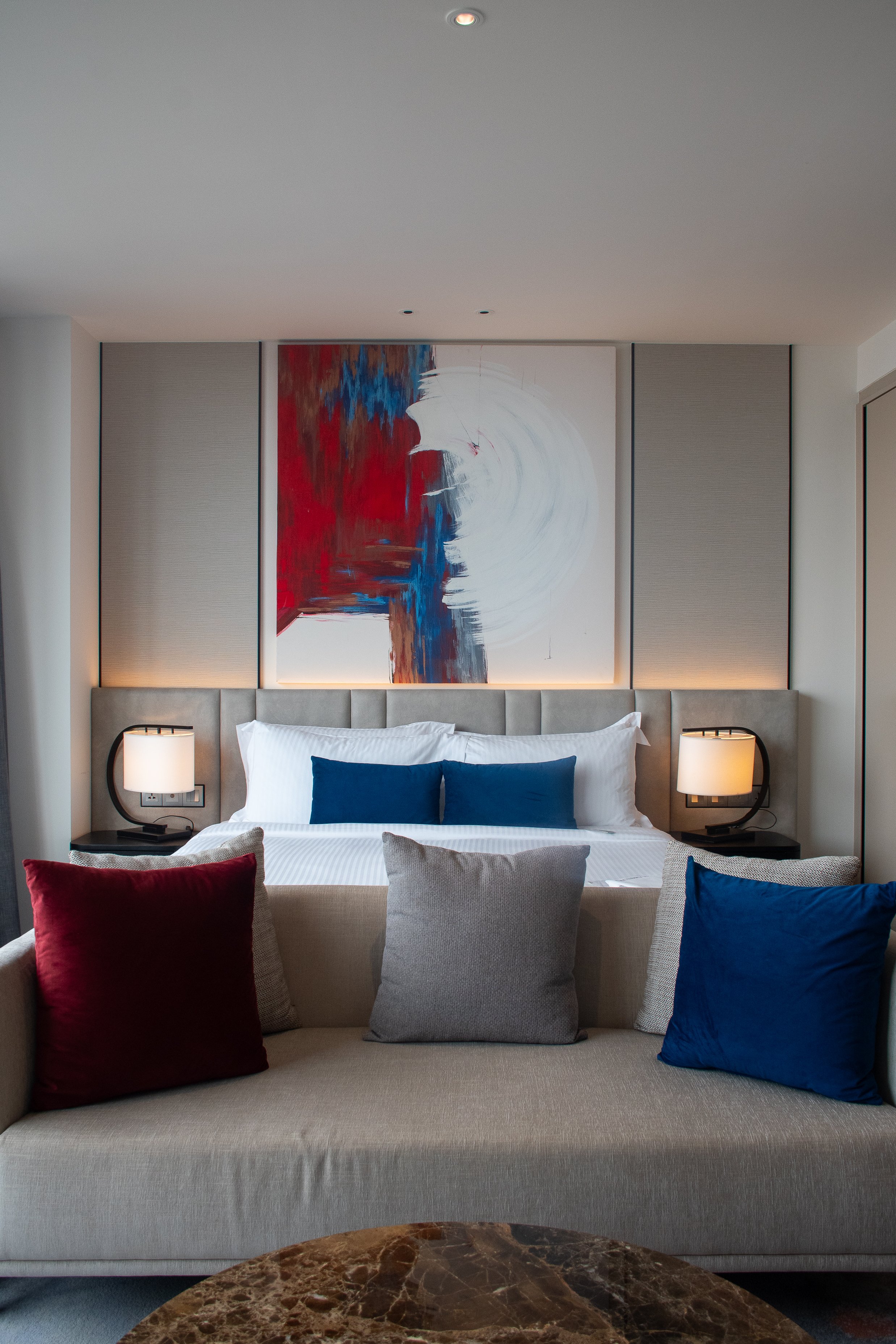 Un lit dans une chambre moderne avec des coussins bleus, gris et bordeaux. Au-dessus du lit, un tableau abstrait avec des couleurs rouge, bleu, blanc et marron. Deux lampes de chaque côté du lit. La chambre a une ambiance élégante et confortable.