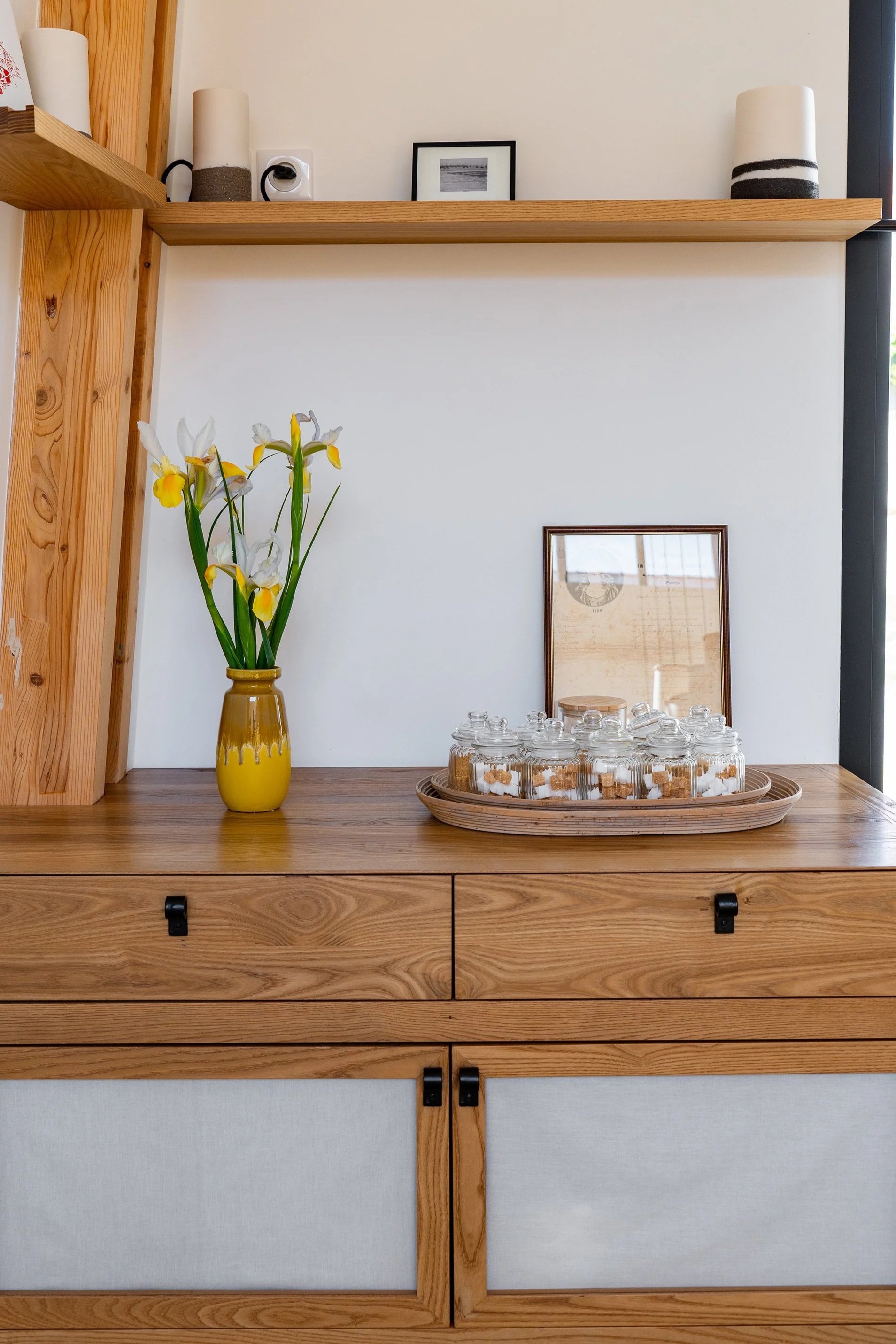 Une étagère en bois avec deux lampes cylindriques, une petite photo encadrée, un vase avec des fleurs blanches et jaunes, et des petits bocaux en verre avec des biscuits, sur un meuble avec deux tiroirs.