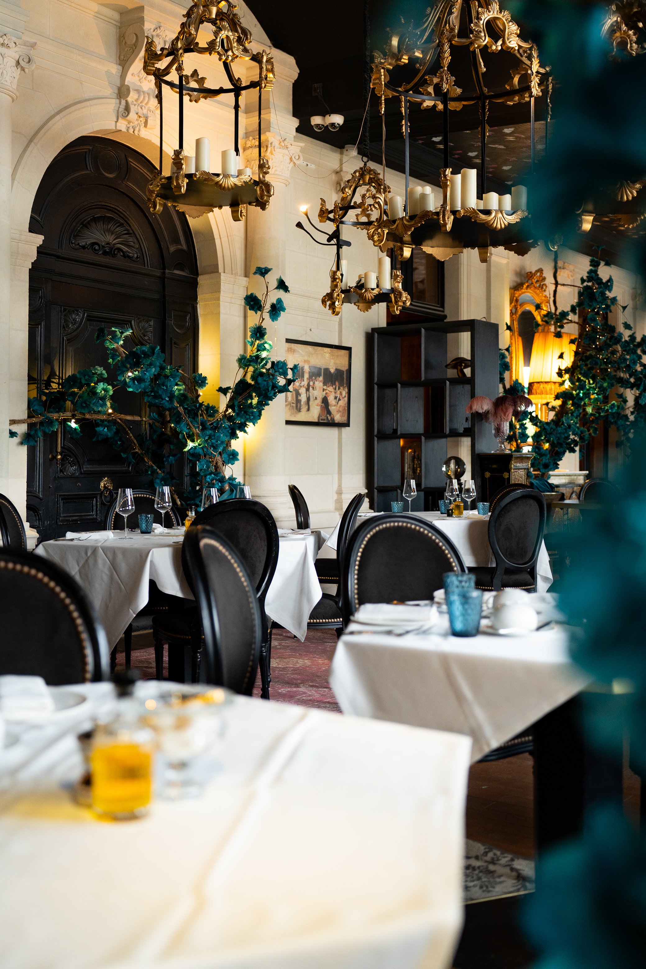 Salle à manger élégante avec des tables dressées, chandeliers dorés suspendus au plafond, décor floral et tableau ornant le mur.