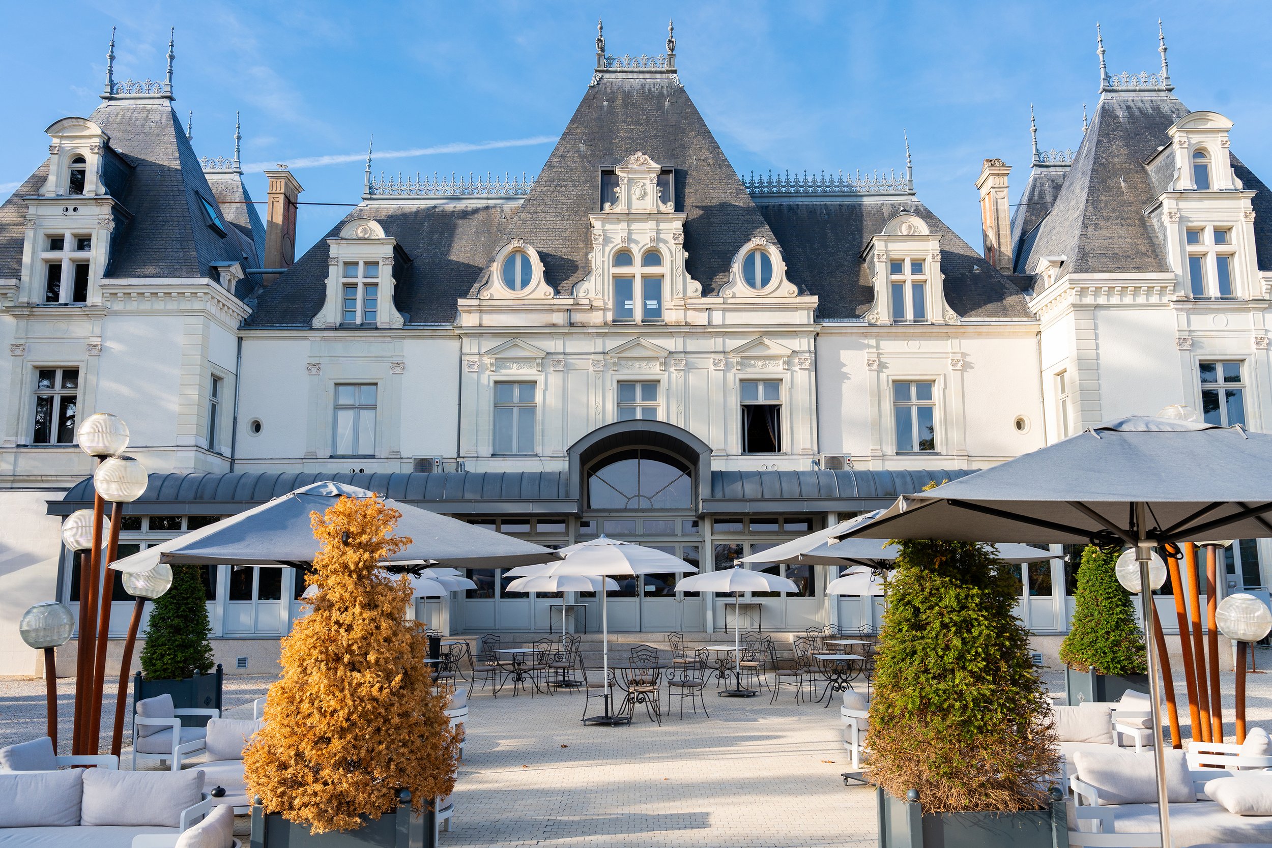 Un bâtiment historique avec une architecture en style Château, toit en ardoise et tourelles, vue arrière avec une terrasse extérieure et des parasols, décorée de buissons et de lampes dans une ambiance de temps clair.