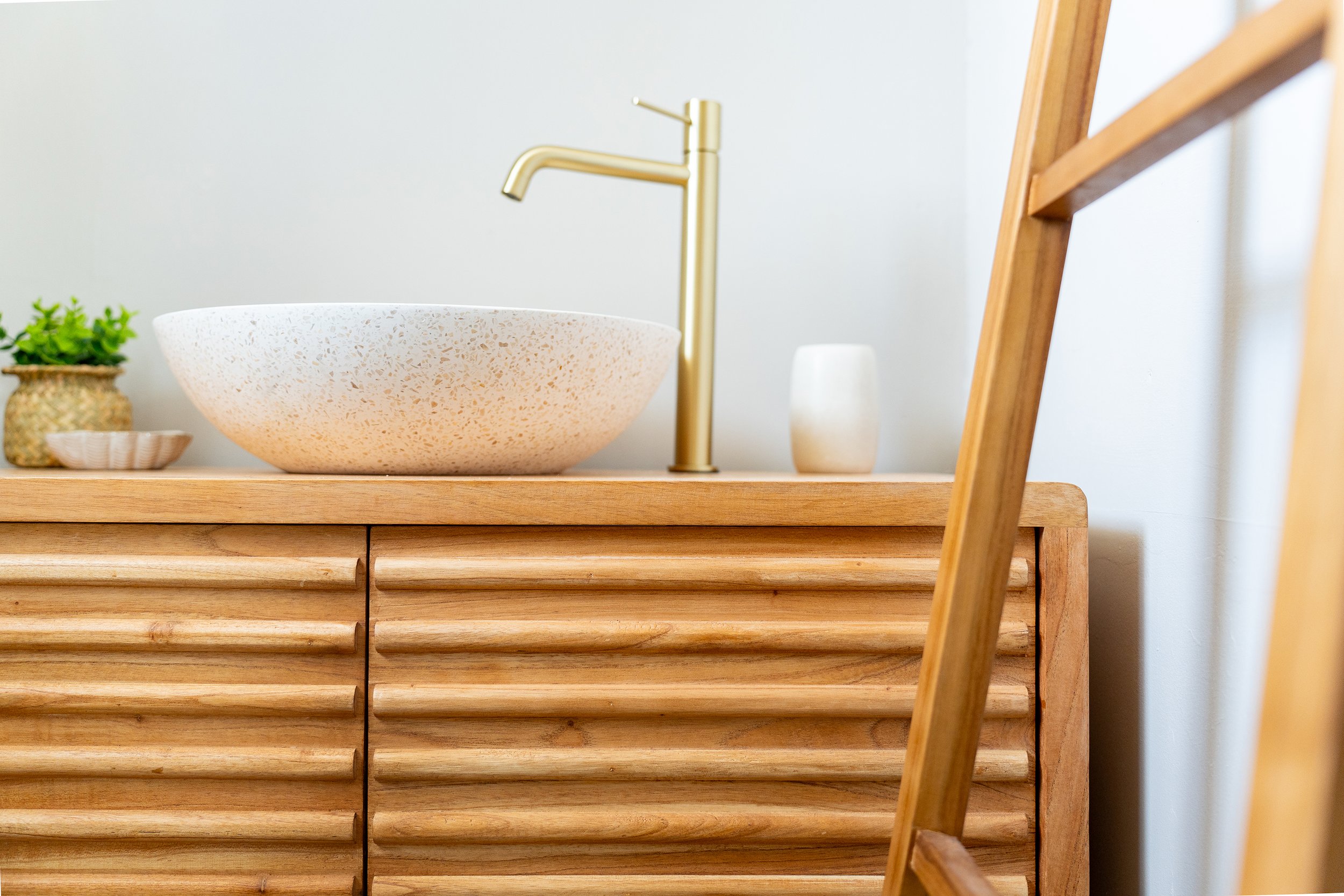 Une salle de bain avec un lavabo en pierre, un robinet en or et une petite plante verte dans un vase en pot. Une chaise en bois est partiellement visible sur le côté droit.