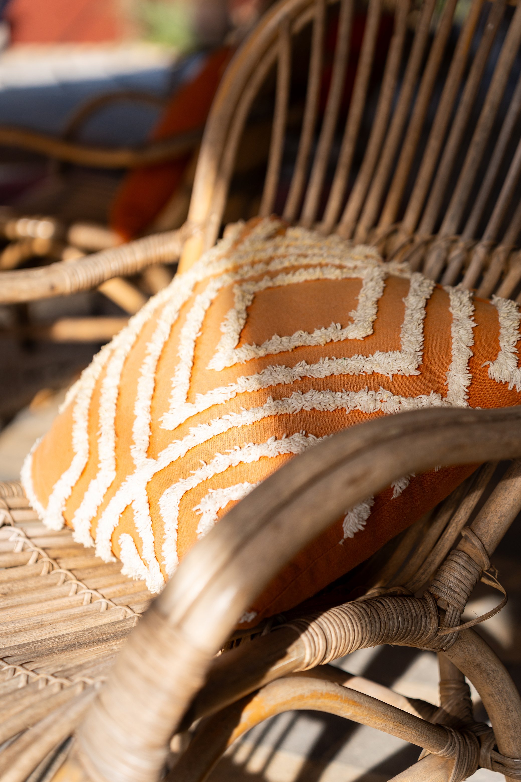 Une chaise en rotin avec un coussin orange décoré de rayures blanches et de motifs en forme de rectangles, prise à l'extérieur sous la lumière du soleil.