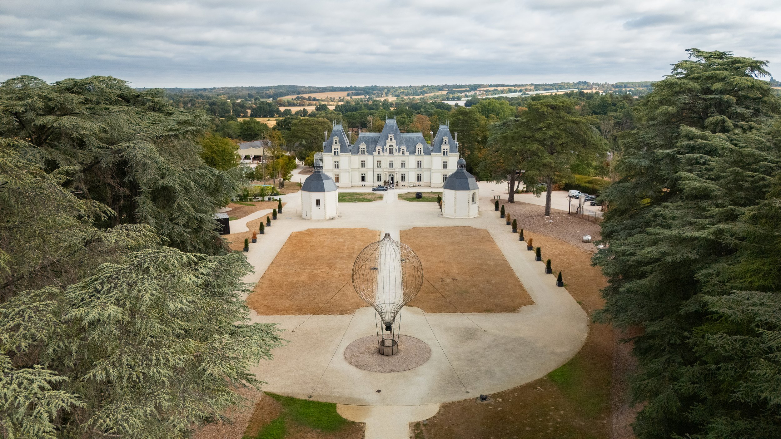 Un château blanc avec des tours et des toits gris, entouré d'arbres et d'un paysage rural, visible d'en haut avec un ciel nuageux.