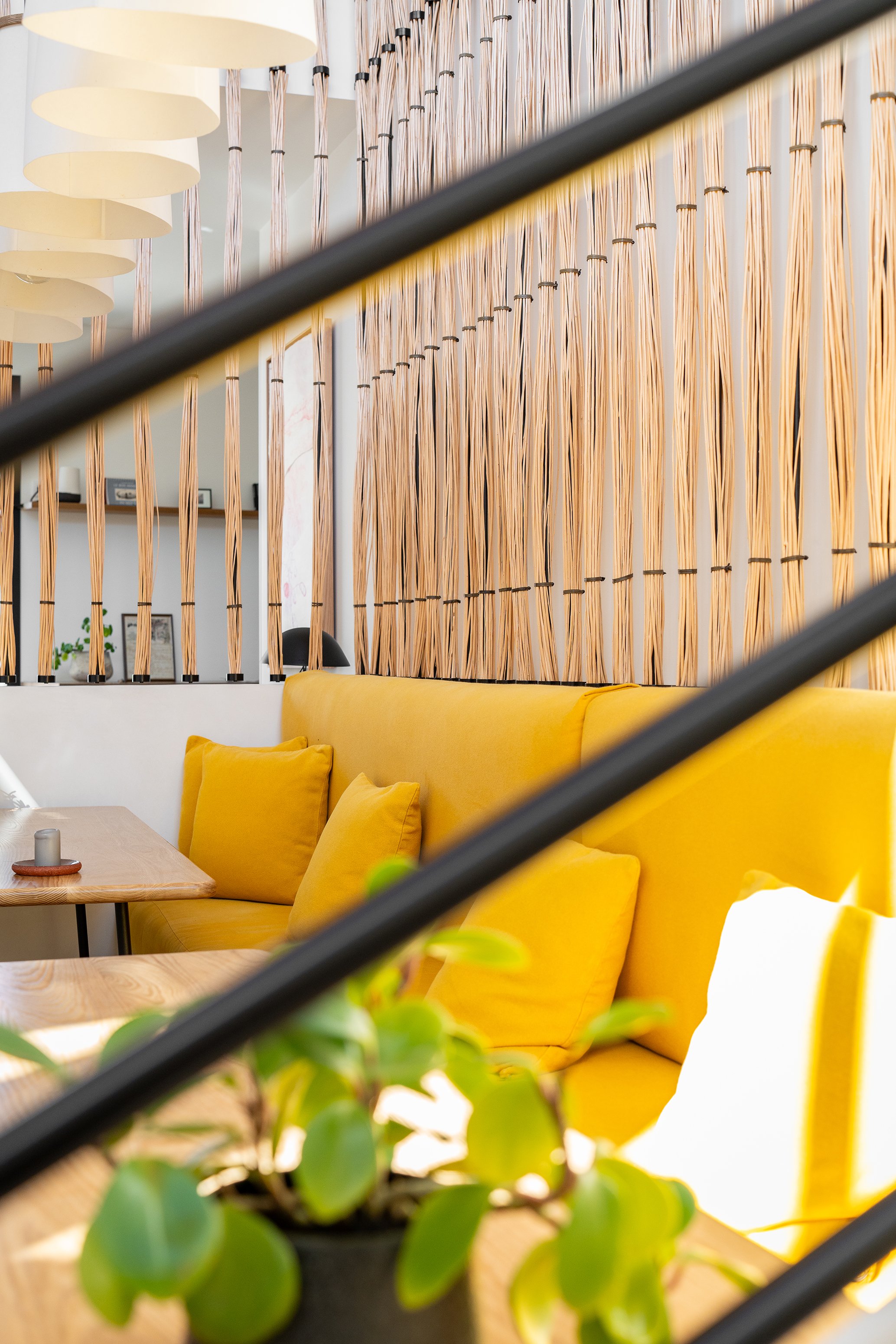 Intérieur d'un espace moderne avec un canapé jaune et des coussins, séparé par une séparation en bois et bambou, décorée avec des plantes vertes, et une lumière suspendue blanche.