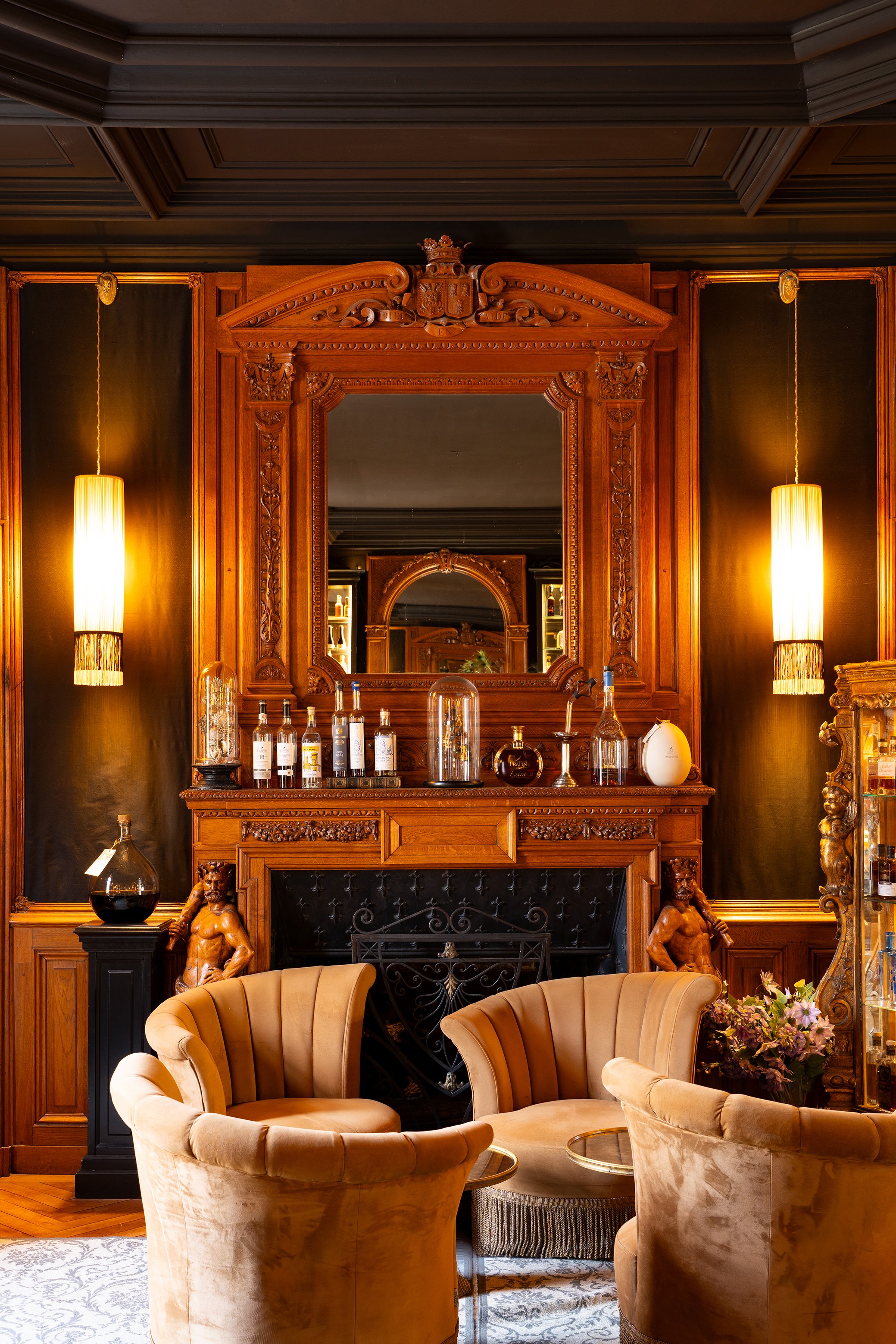 Intérieur d'une salle élégante avec cheminée en bois sculpté, miroirs encadrés en bois, fauteuils en velours beige,-luminaires suspendus, objets décoratifs dont bouteilles et statues, fleurs dans un vase.