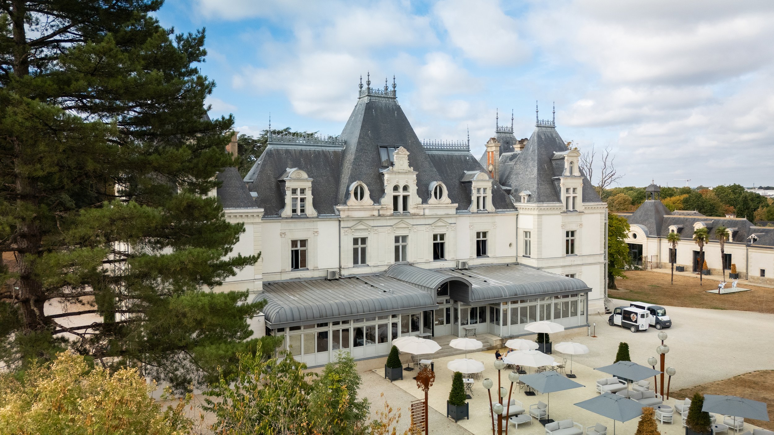 Un grand manoir blanc avec un toit en ardoise, entouré d'arbres et d'un espace extérieur avec des parasols blancs et des meubles, sous un ciel partiellement nuageux.