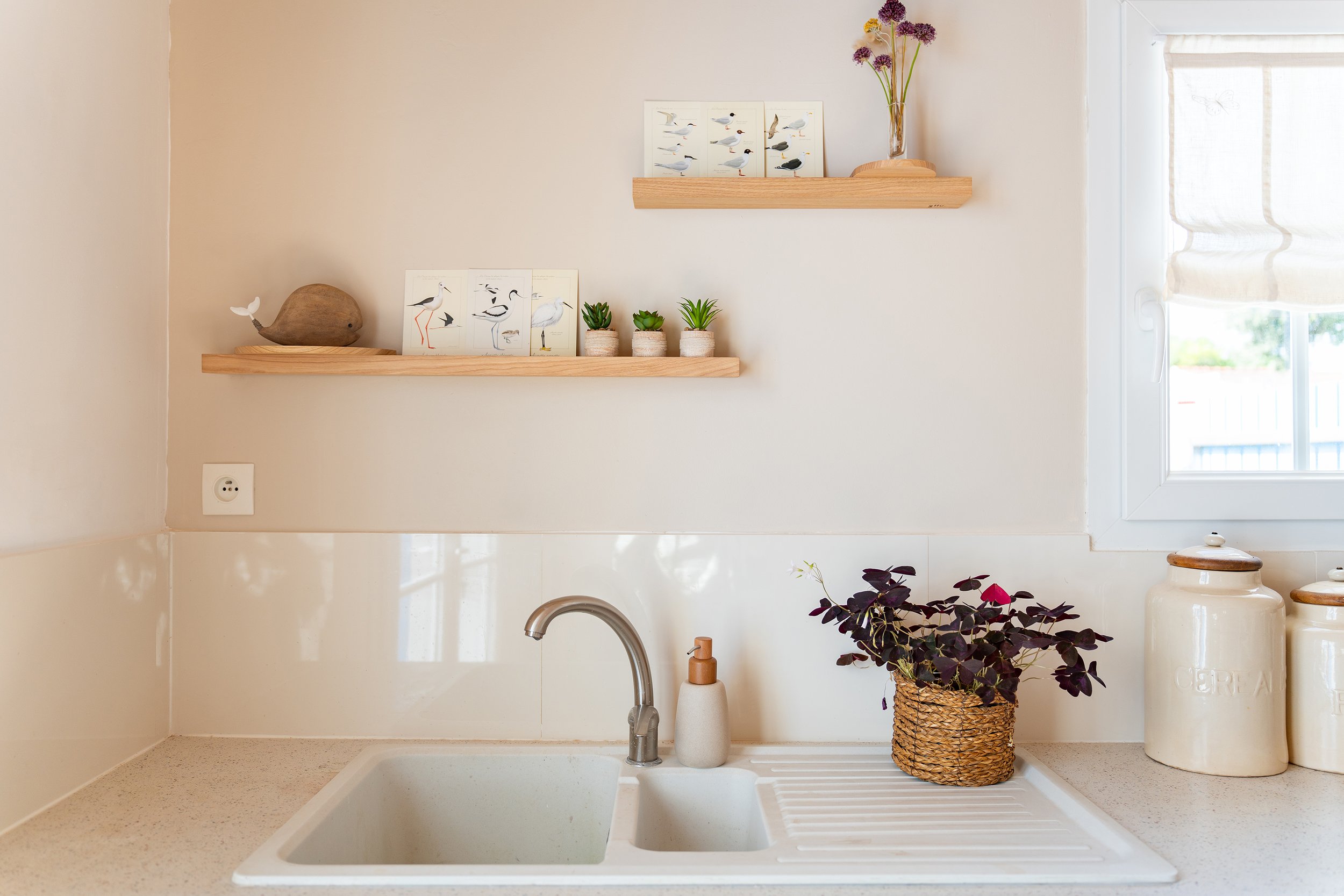 Évier de cuisine avec une plante en panier en osier et deux vases blancs, sous deux étagères en bois ornées de décorations de oiseaux et de fleurs violettes, avec une fenêtre à droite et une prise électrique.