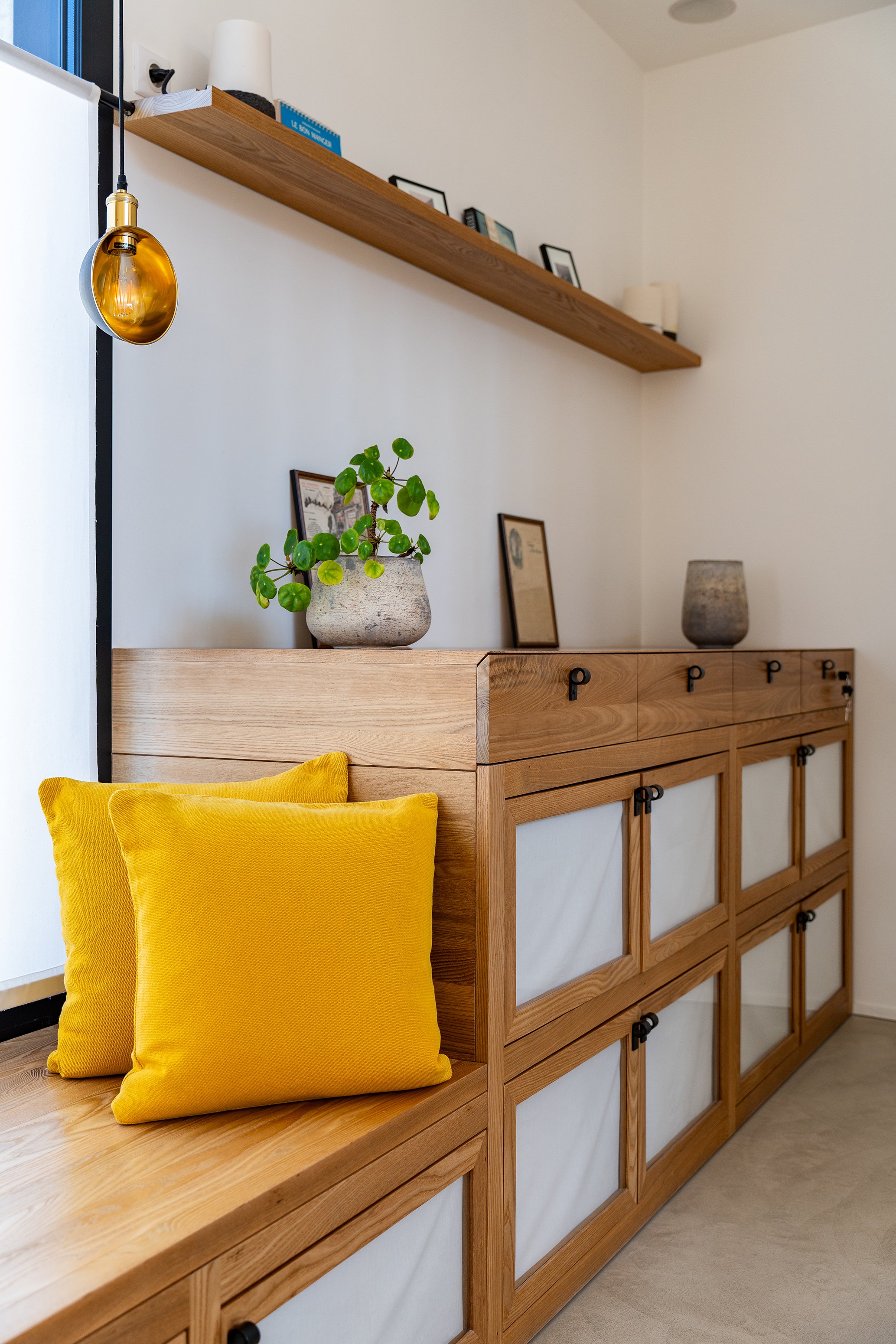 Une commode en bois avec des tiroirs en bois clair et des panneaux en crépi blanc, decorée avec deux coussins jaunes en velours et une plante verte en pot.
