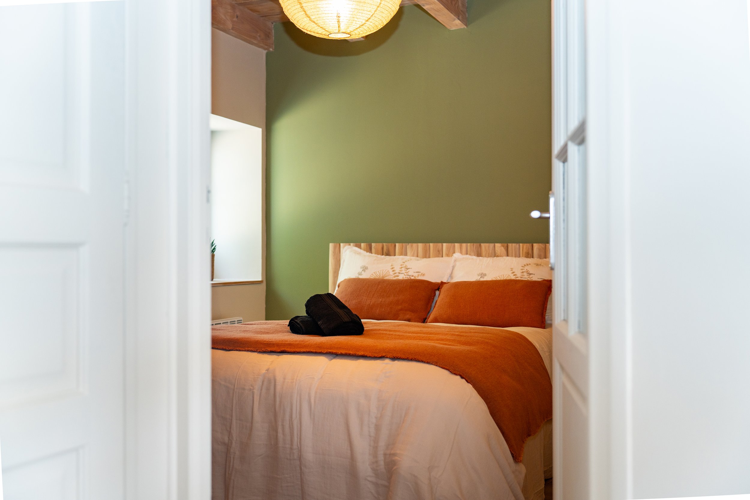L'image montre une chambre vue à travers une porte entrouverte. La chambre a un mur vert, un lit avec une tête de lit en bois, une couette beige et des coussins marron. Il y a un lampadaire en papier au plafond.