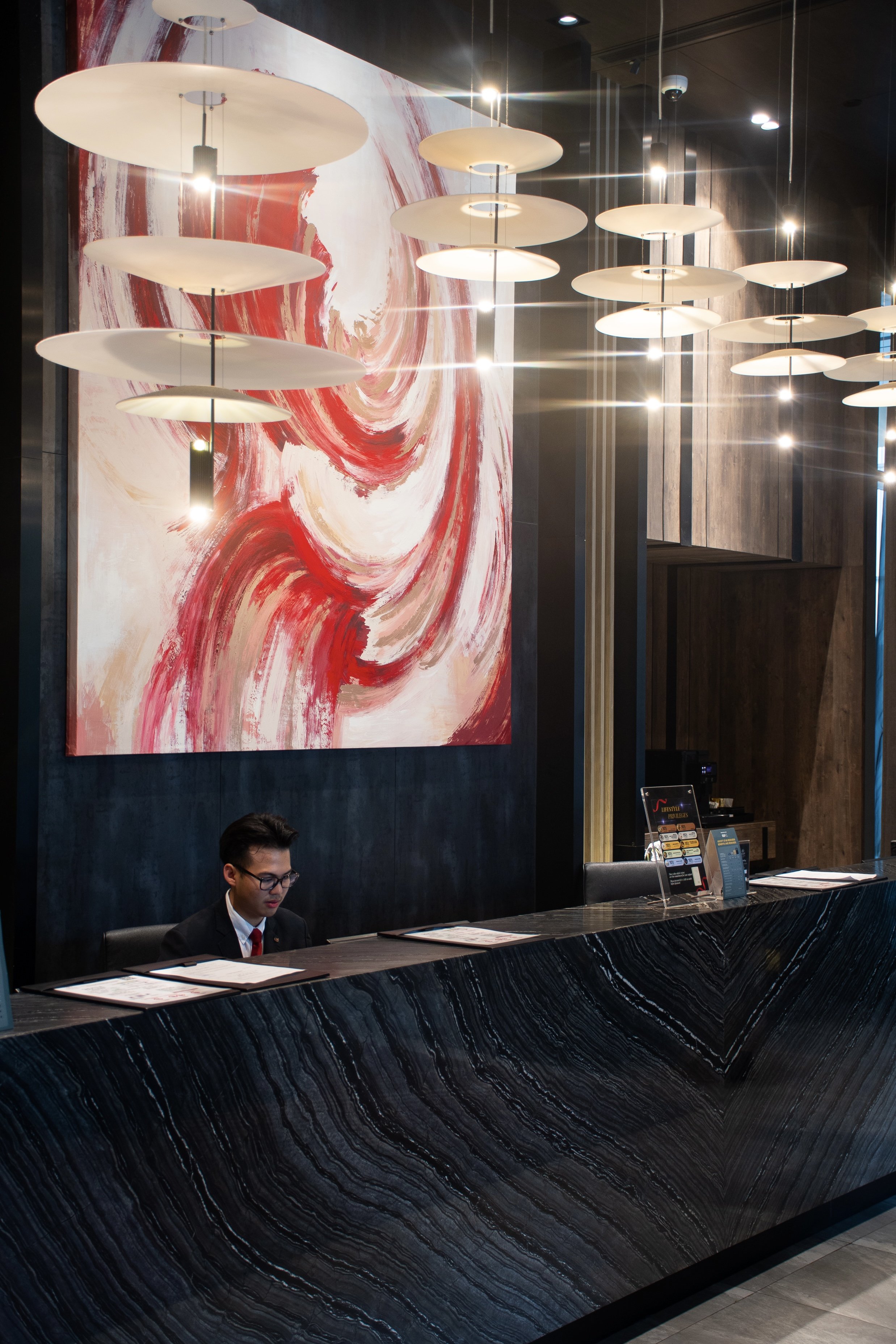 Un guichet d'hôtel avec un homme assis derrière le comptoir en marbre noir, un tableau abstrait avec des couleurs rouge, blanc et beige suspendu au mur derrière lui, et une table avec des panneaux d'information sur le comptoir.