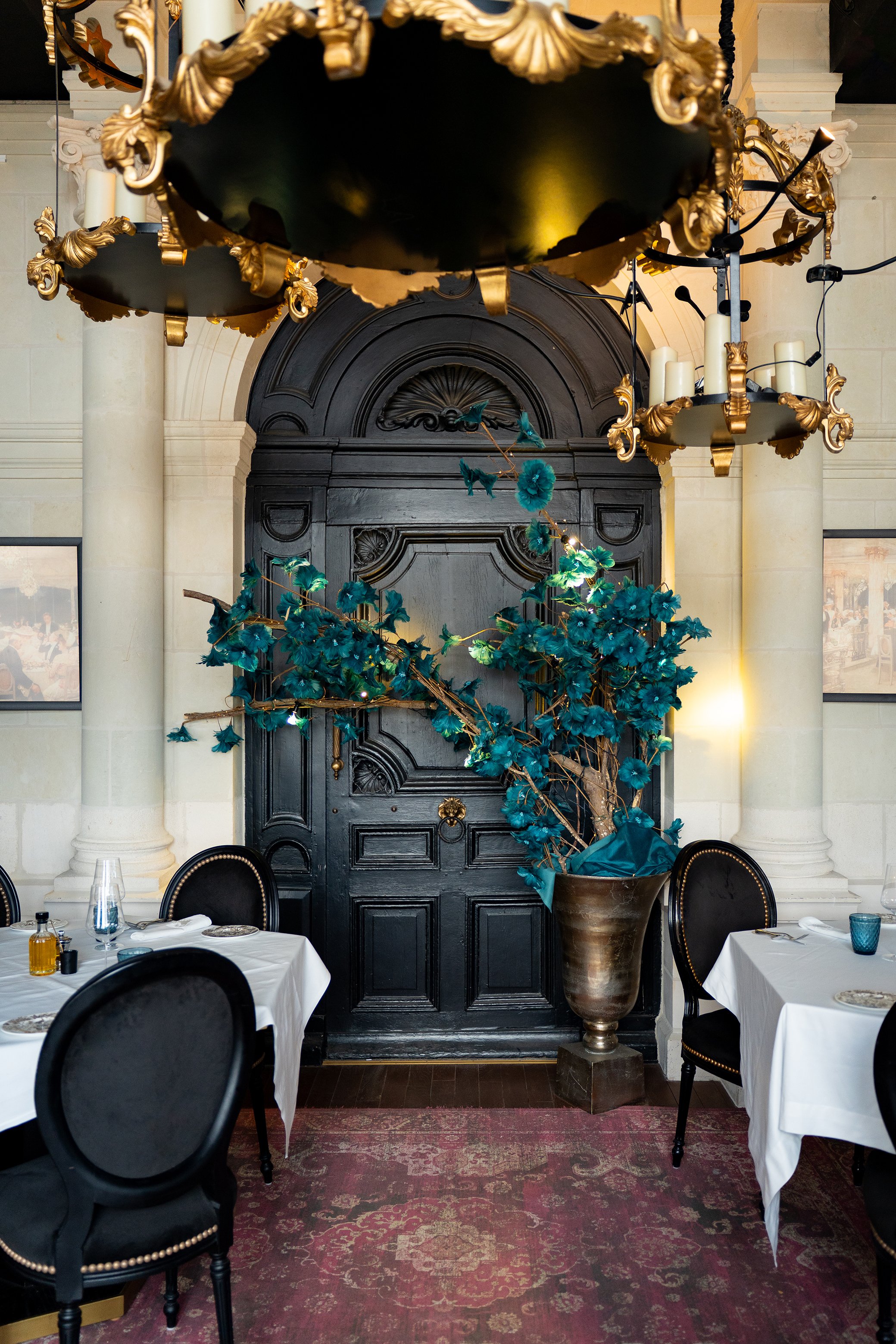 Intérieur d'un restaurant élégant avec des tables dressées pour un repas, des chaises noires, une grande poterie avec une plante décorative et des lustres dorés.