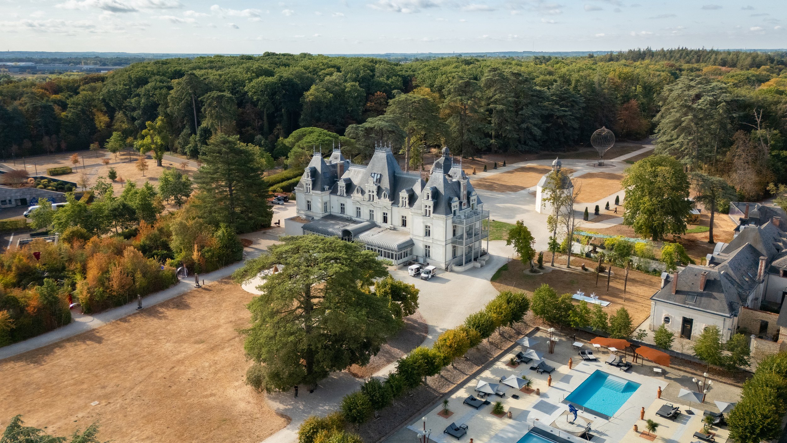 Vue aérienne d'un château blanc avec plusieurs toits pointus, entouré de grands arbres et d'un parc.
