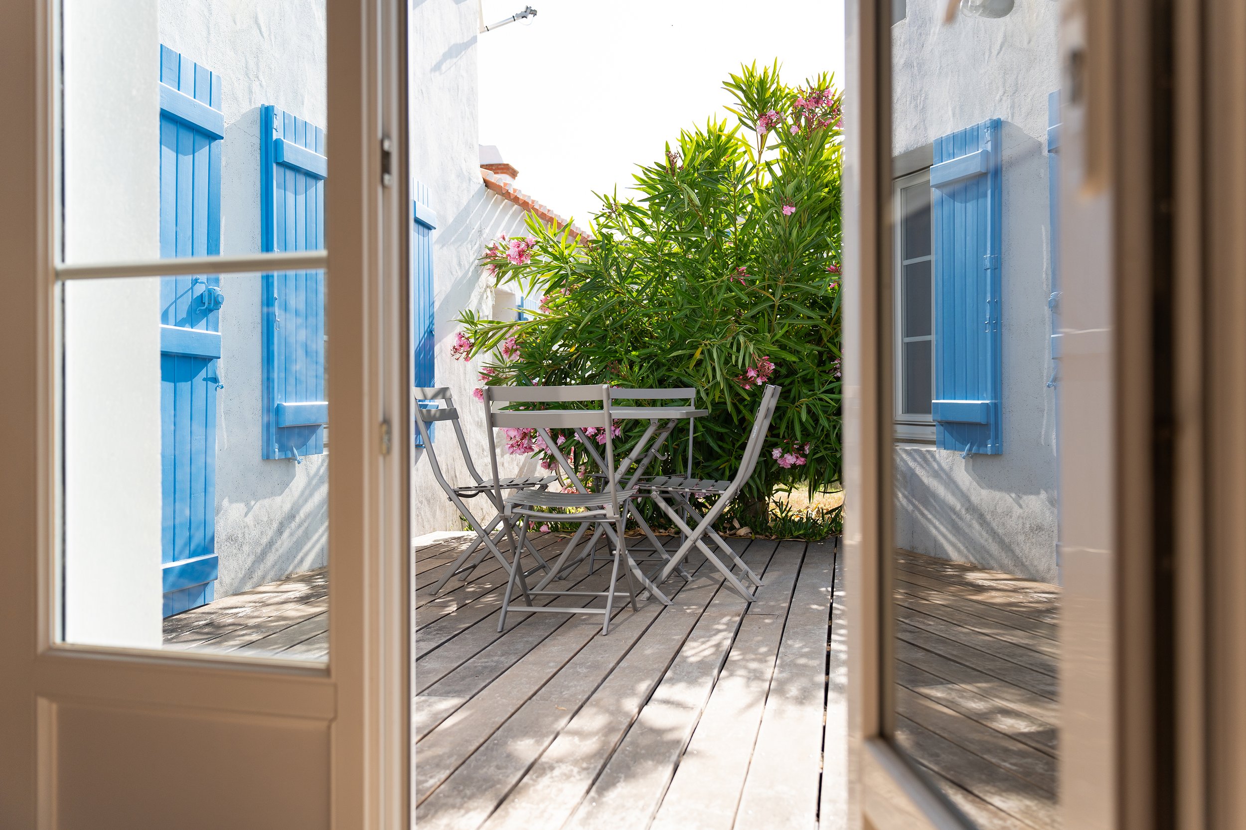 Vue d'une terrasse avec une table et des chaises en métal, entourée de plantes avec des fleurs roses et un mur blanc avec volets bleus, vue à travers une fenêtre.