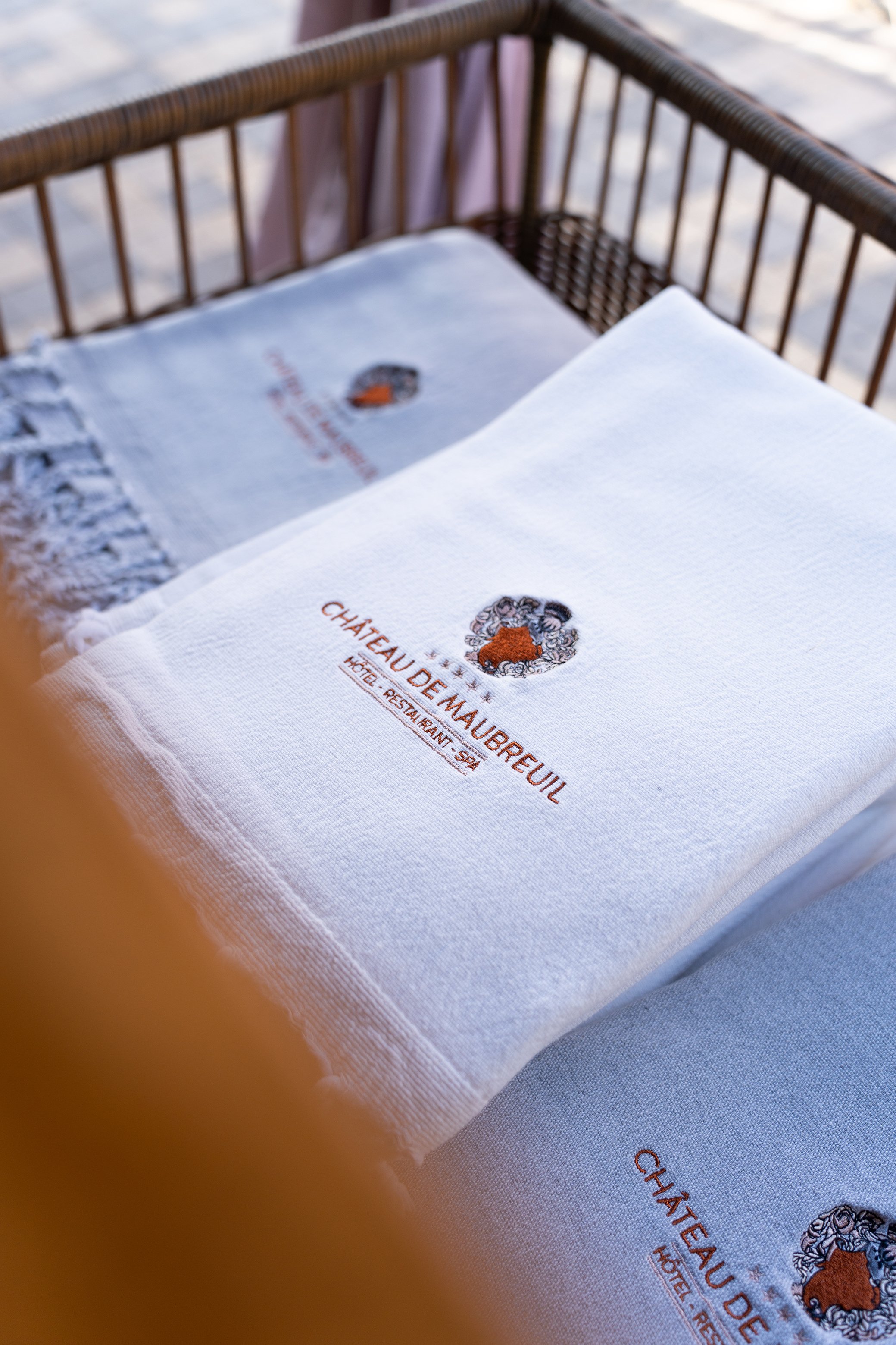 Serviette blanche avec le logo du Château de Maubreuil, un château entouré d'un cercle de fleurs, posé sur une table à l'extérieur, avec un fond flou de sable ou de plage.