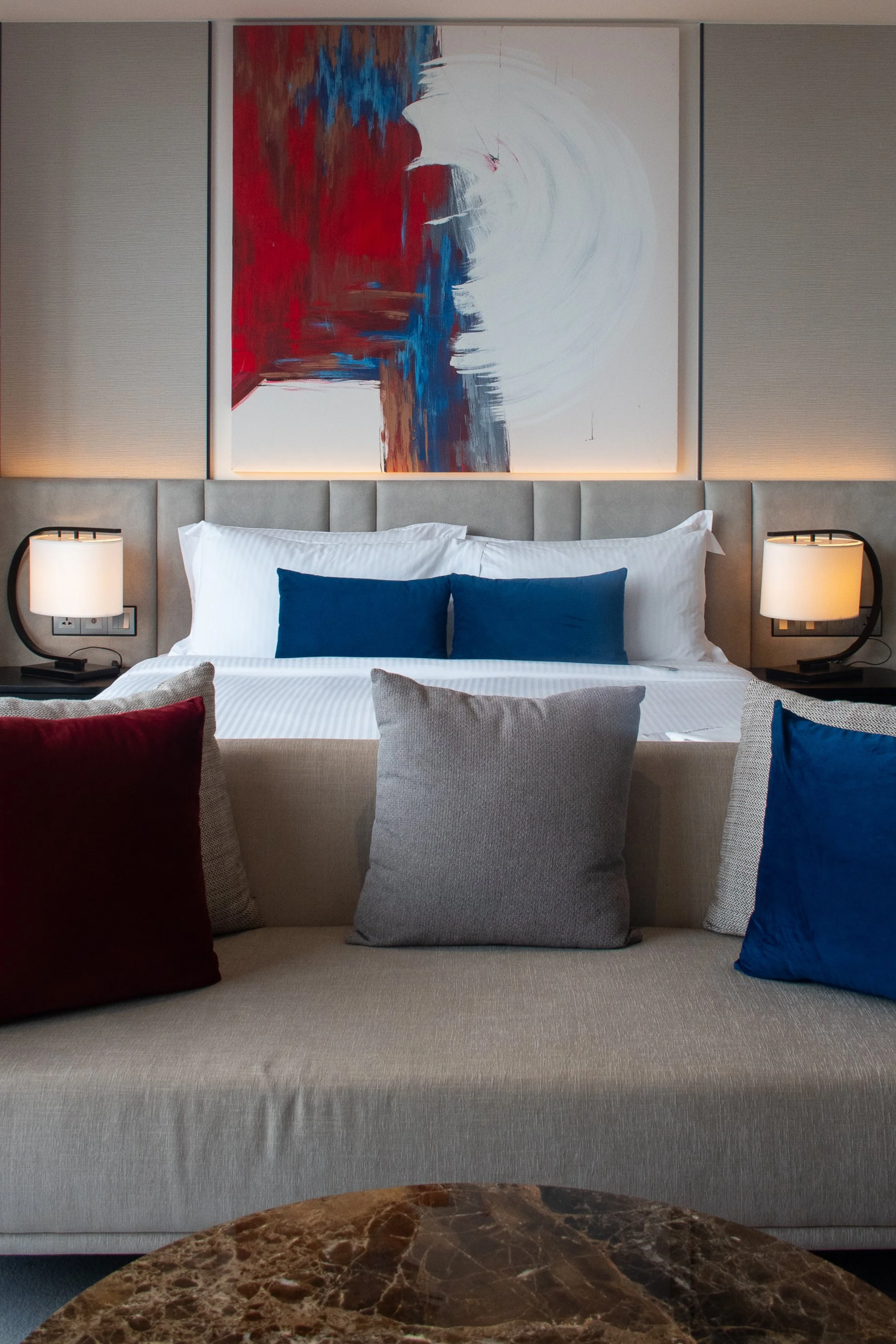 Un lit avec des oreillers blancs et deux coussins bleus, devant un tableau abstrait. Cette chambre est accueillante avec deux lampes de chaque côté du lit. Un canapé coloré est visible au premier plan.