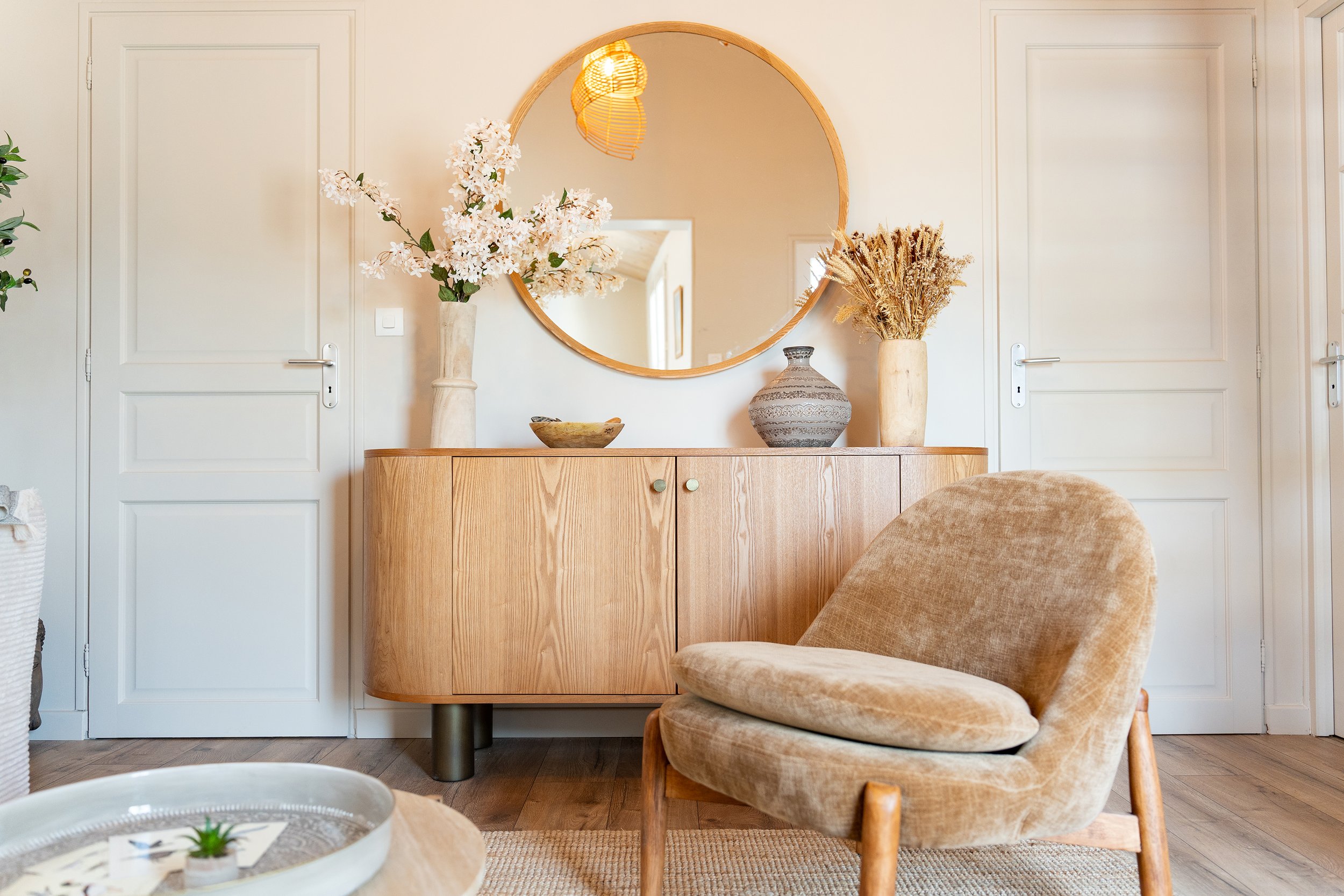 Intérieur d'une pièce avec un meuble en bois, un miroir rond, des vases avec des fleurs séchées et peu de décoration, avec deux portes blanches en arrière-plan.