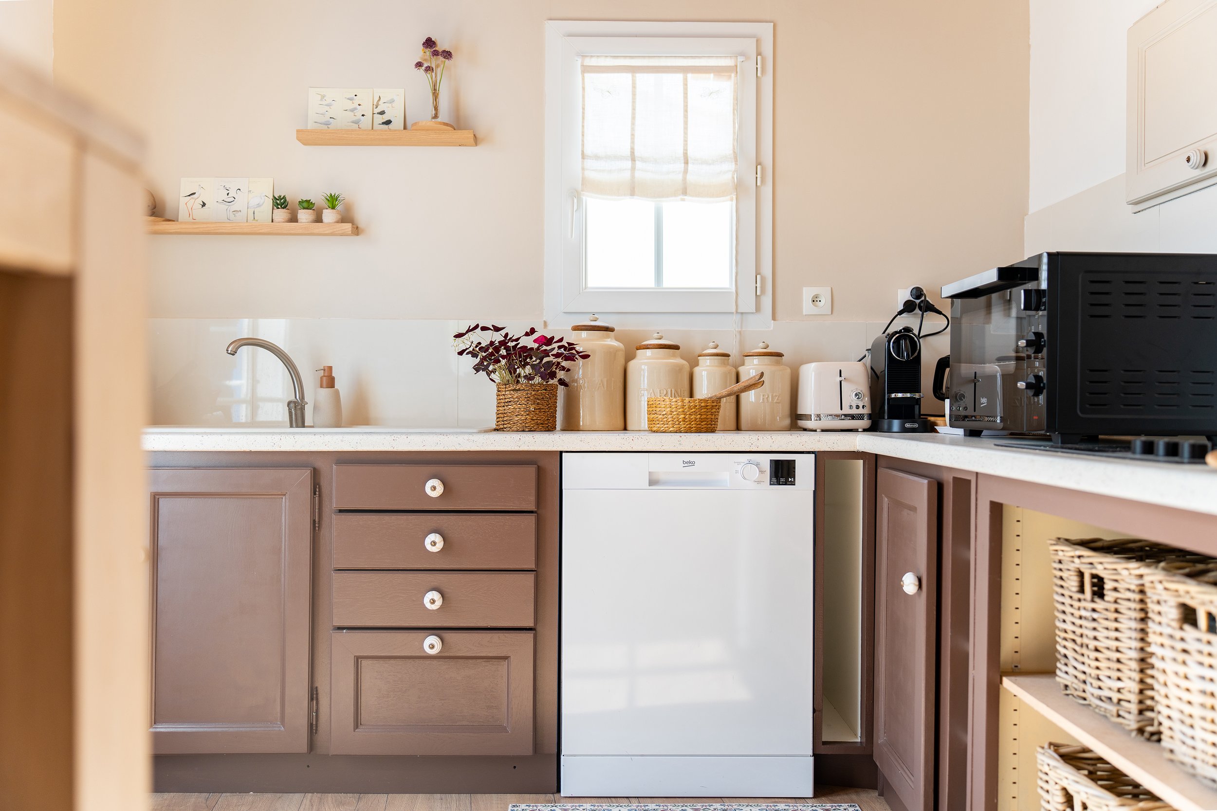 Cuisine propre avec un évier, des pots en céramique, une plante, une machine à café, un grille-pain, un four micro-ondes, une fenêtre avec rideau beige, et des étagères en bois avec des décorations de oiseaux.