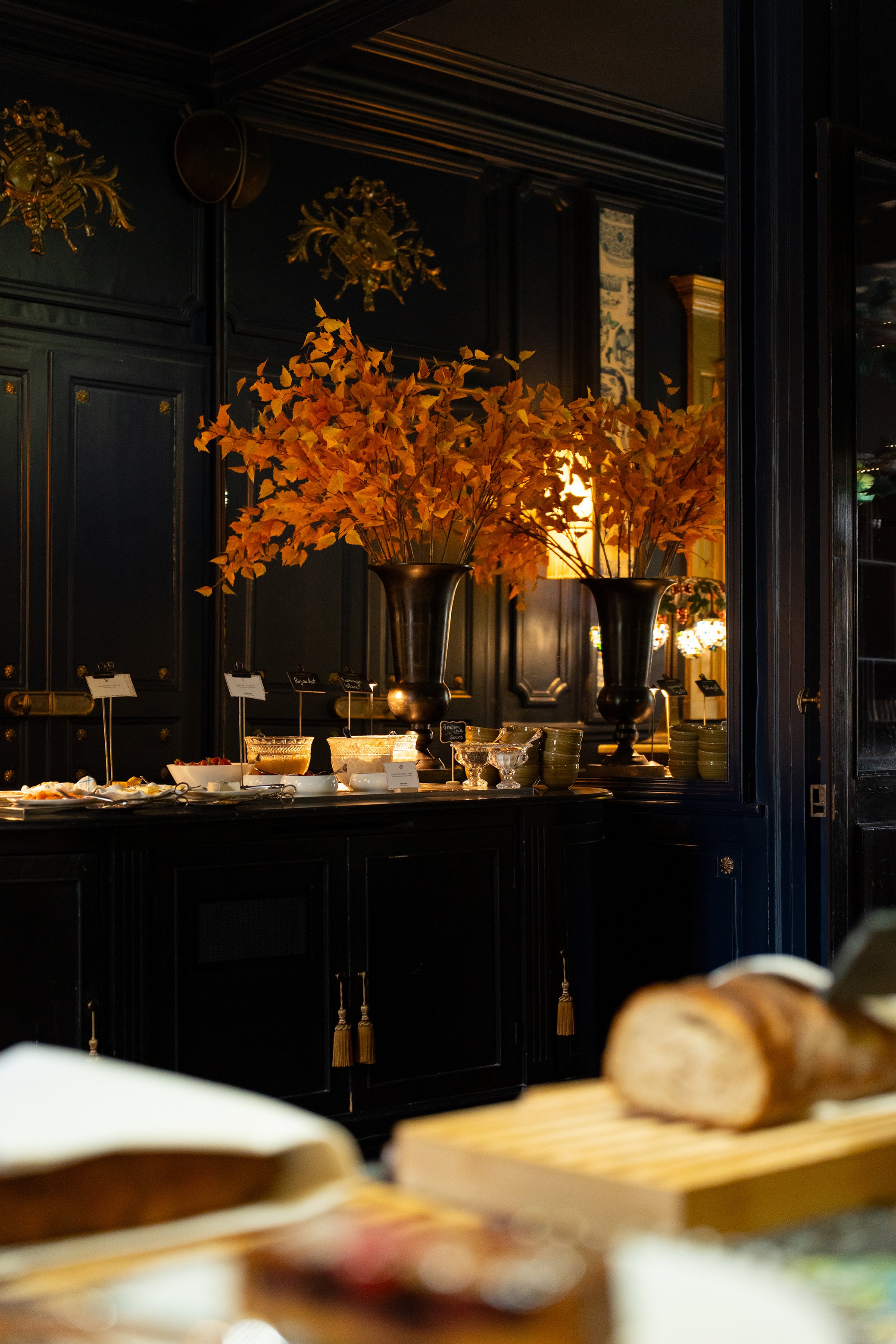 Vases avec des branches de feuilles dorées dans un décor élégant avec des murs noirs et des luminaires en or, ambiance chaleureuse et sophistiquée.