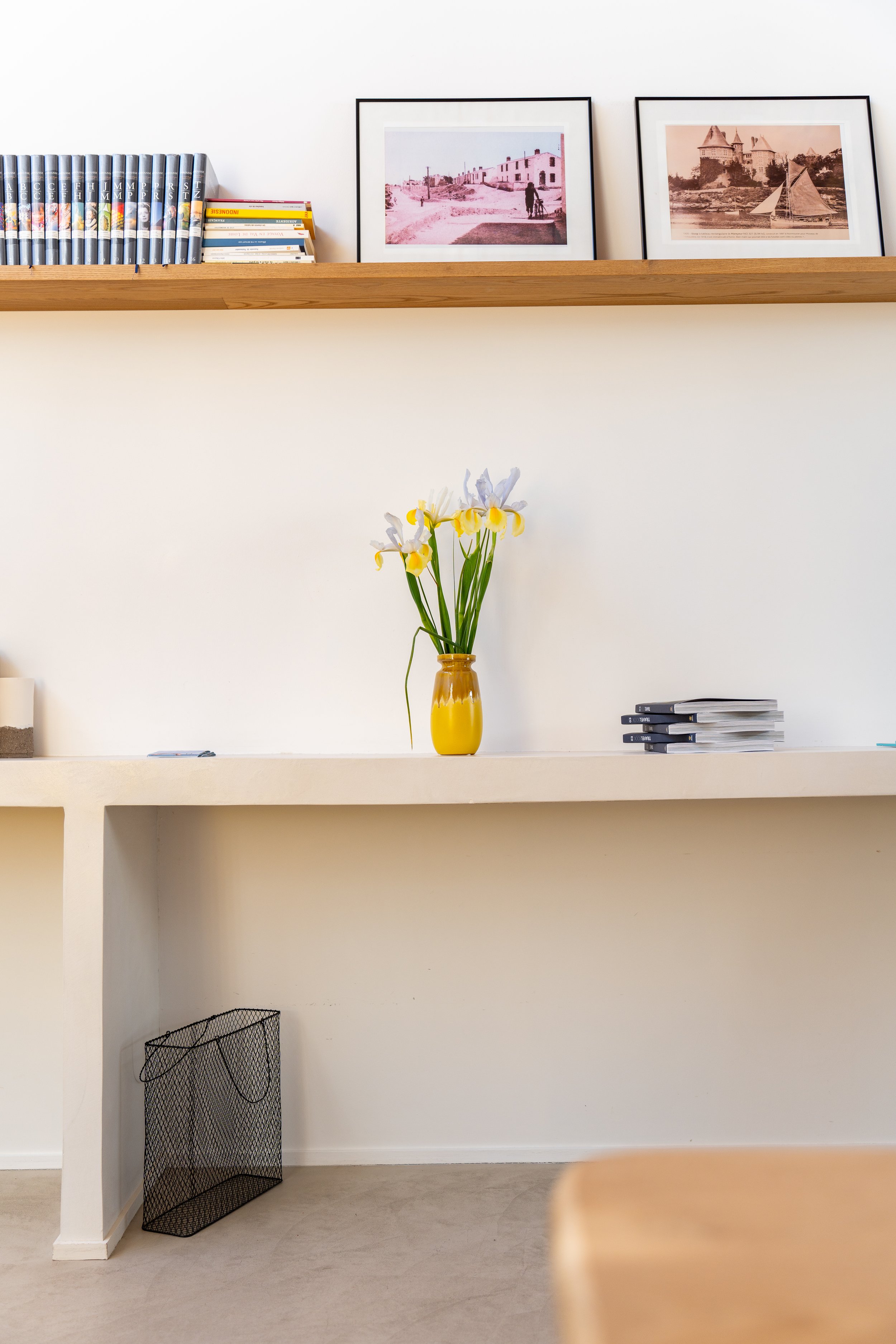 Une étagère en bois avec deux cadres photo, une pile de livres, une vase jaune avec des fleurs, un panier à la base, et un mur blanc.