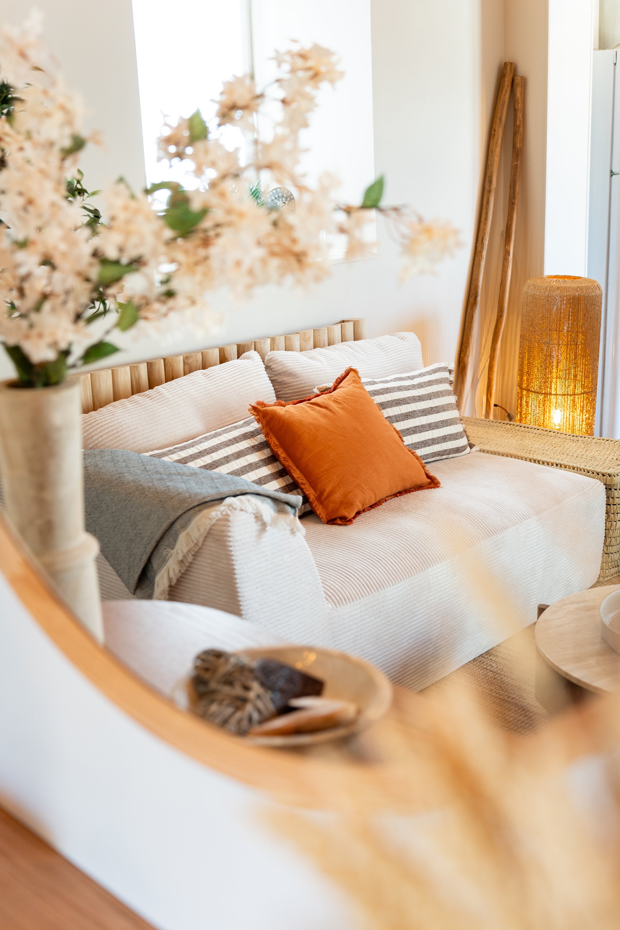 Un coin salon avec un canapé beige, décoré avec des coussins orange et rayés, une lampe en osier, et une plante avec des fleurs blanches en premier plan.