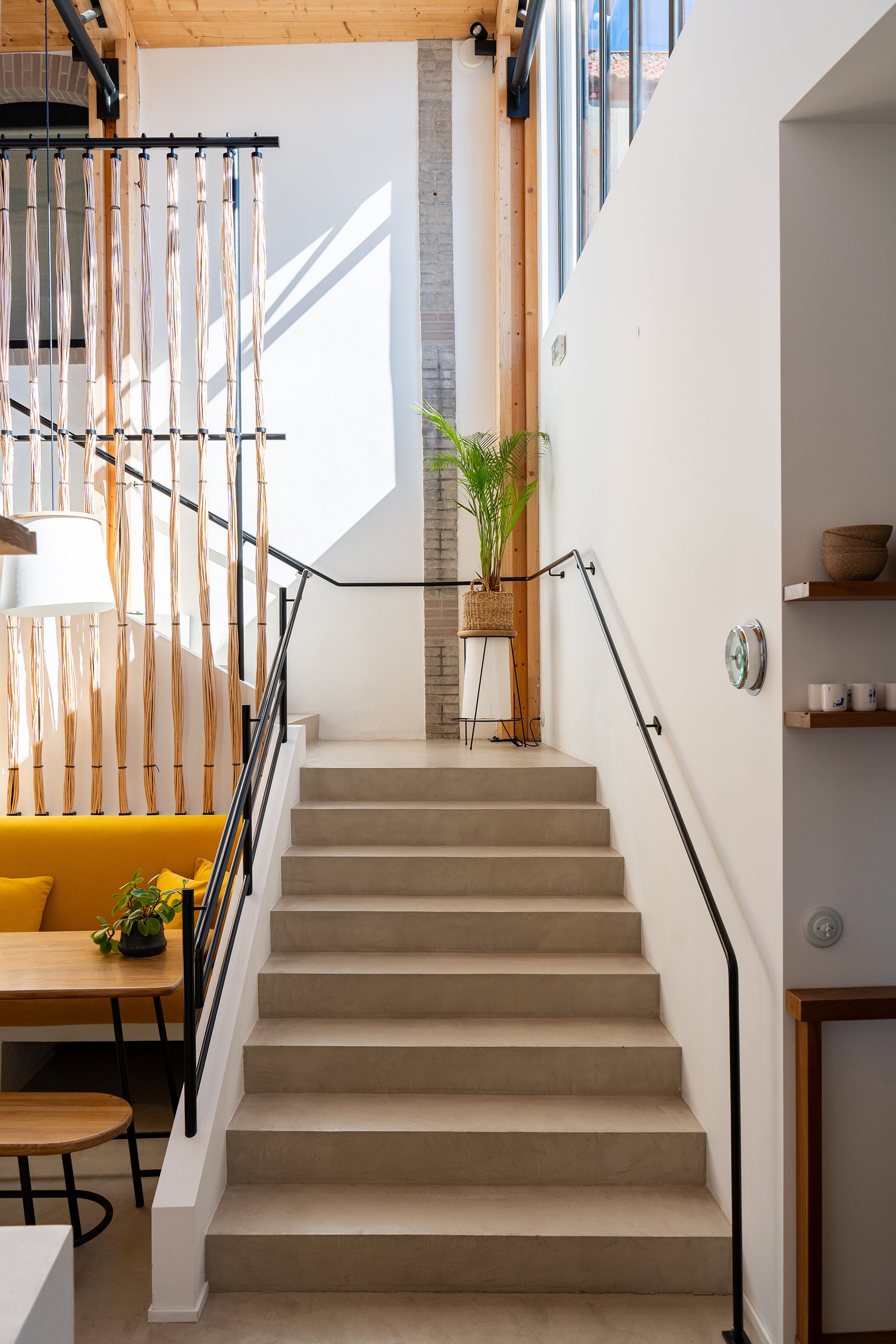 Escalier intérieur en béton avec des rampes en métal noir, une plante verte en pot en osier, et une grande fenêtre laissant entrer la lumière naturelle. Mur blanc avec une étagère en bois contenant des céramiques et une horloge