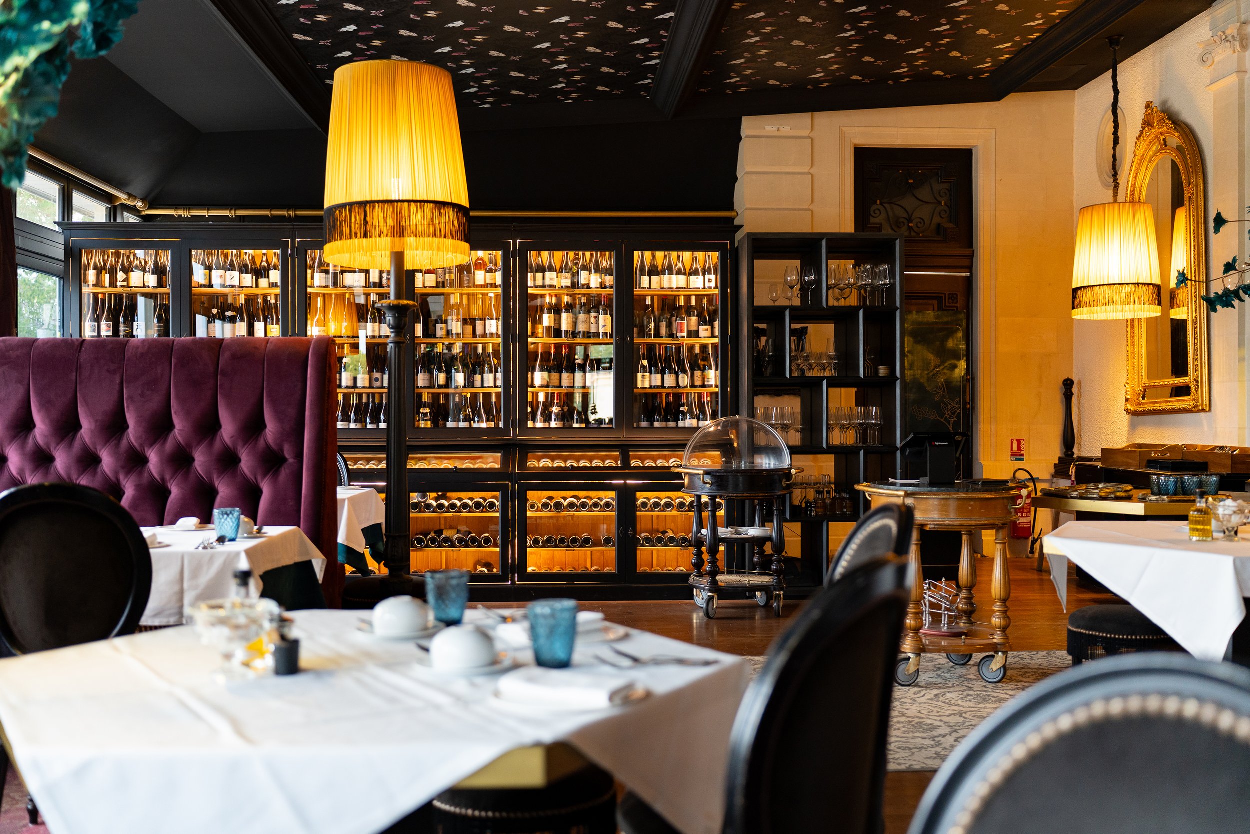 Restaurant élégant avec des tables ornées de nappes blanches, des chaises en velours violet, des lampes jaunes et des étagères à vin en arrière-plan.