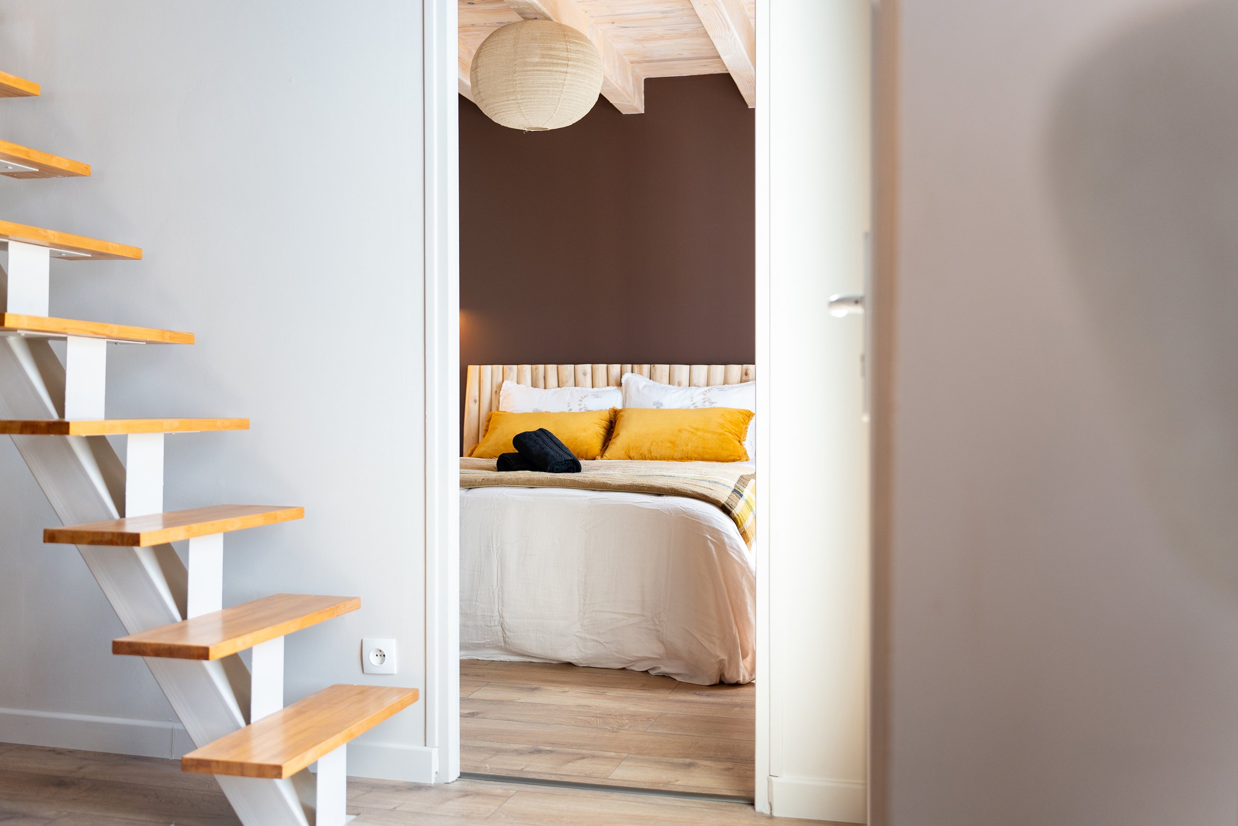 Vue partielle d'une chambre avec un lit dont la tête de lit en bois, des coussins jaunes et un plaid à carreaux. Une lampe en papier suspendue au plafond et un mur marron en arrière-plan. Le point de vue est à travers une ouverture, avec une échelle 