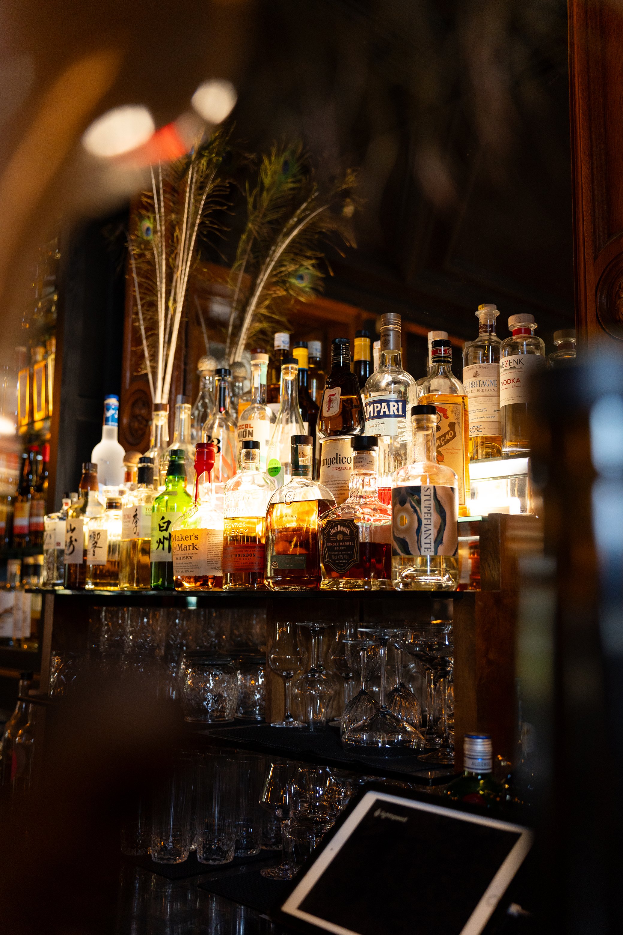 Etal de bouteilles d'alcool derrière le bar avec des verres en dessous, décorations de plumes de paon en haut à gauche.