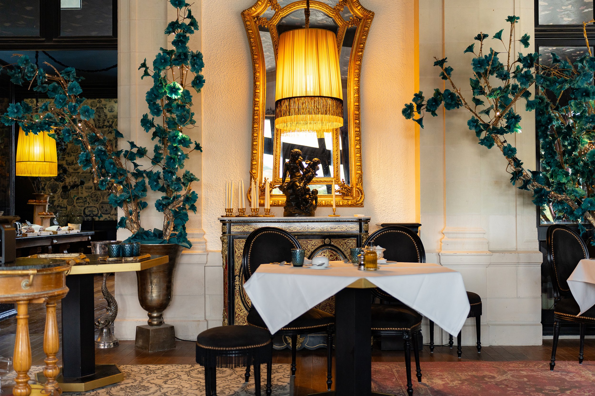 Intérieur d'un restaurant élégant avec un miroir doré, un lustre avec un abat-jour jaune, des décorations florales en bleu et une table avec un napperon blanc, des chandeliers et des assiettes.