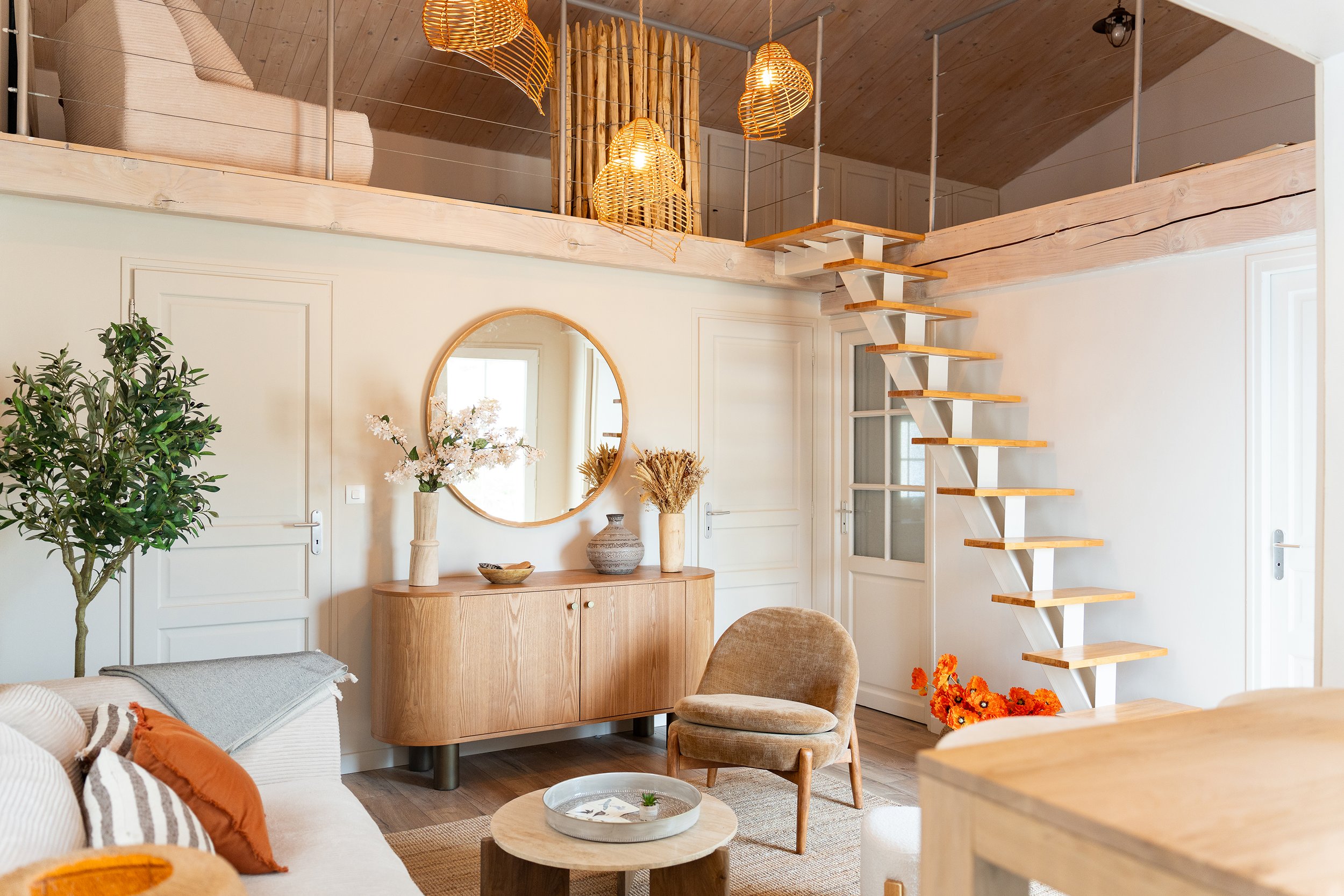 Intérieur d'un salon avec canapé beige, table basse en bois, fauteuil en tissu beige, portes blanches, miroir rond, table basse, et décorations florales, avec un escalier en bois menant à un loft suspendu