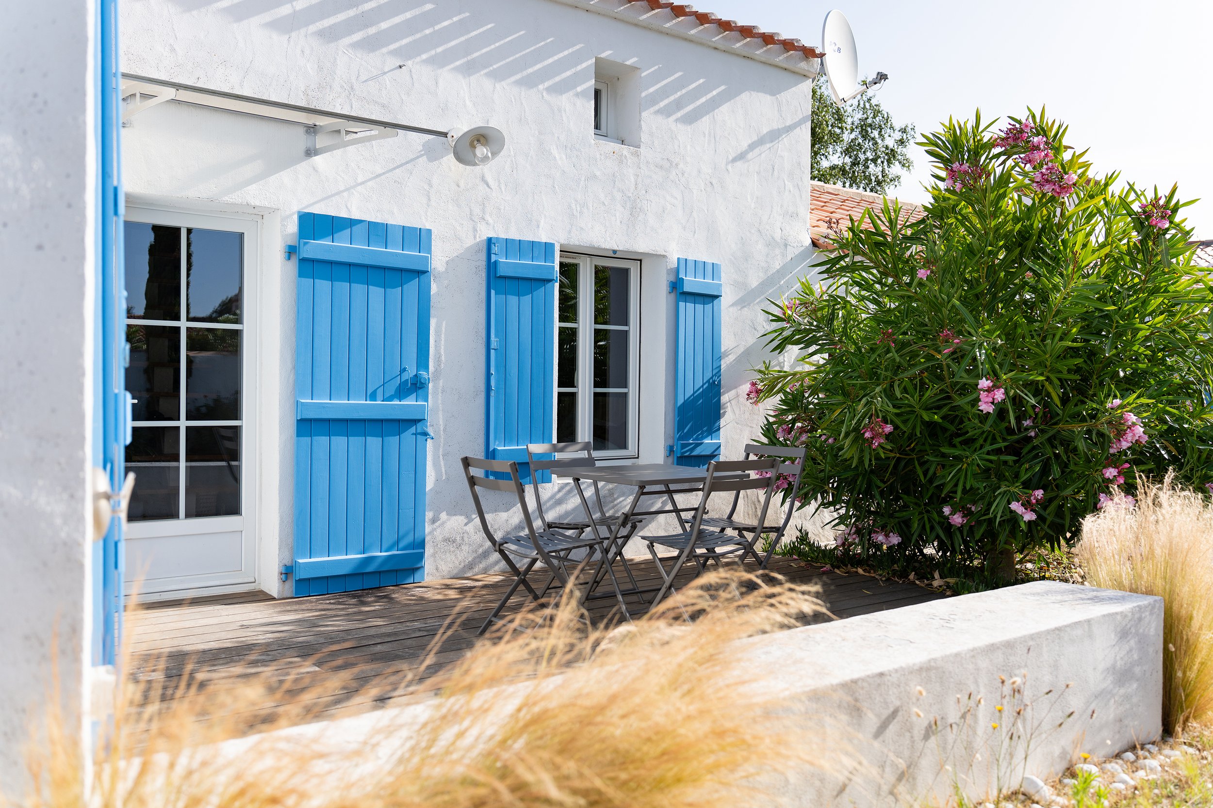 Facade blanche d'une maison avec volets bleus, une porte vitrée, une table en métal avec deux chaises en bois, et un grand arbuste fleuri avec des feuilles vertes et des fleurs roses. Il y a aussi un satellite et un éclairage extérieur.