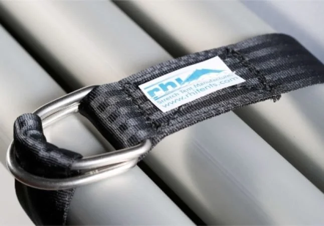 Close-up of a black strap with a metal loop and a label, attached to aluminium poles,  likely a trailer or cargo strap.
