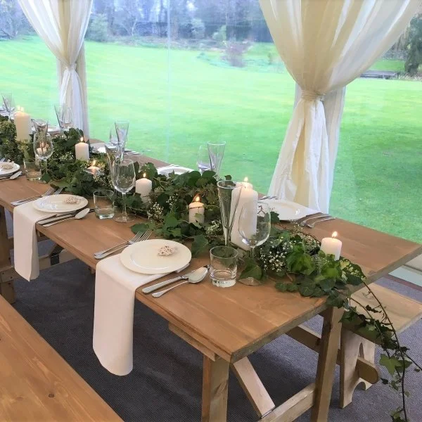 Decorated dining table with candles, greenery, and glassware in a bright room with large windows and white curtains, set for a meal.