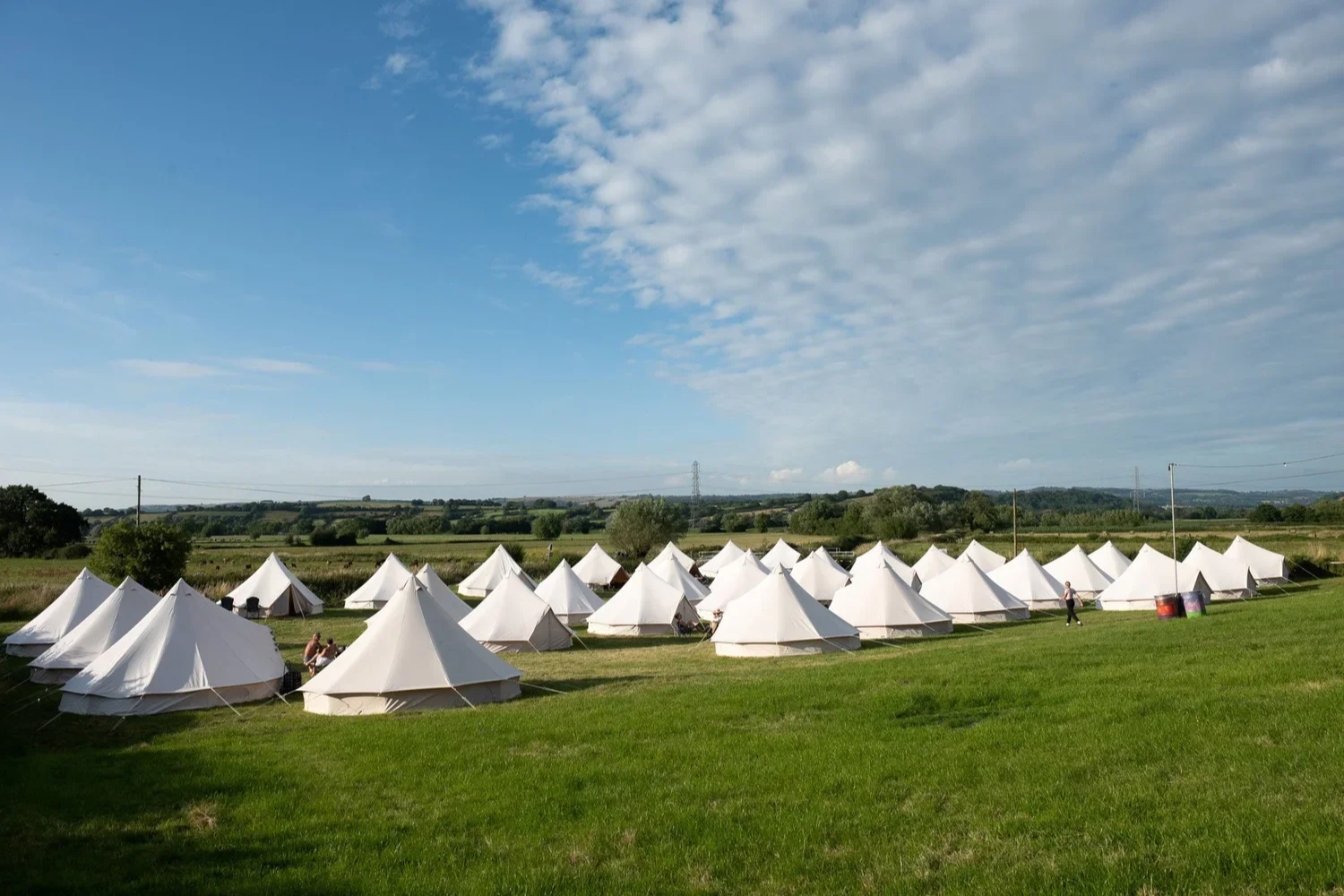 A field with numerous white canvas tents set up in rows on a lush green lawn, with a blue sky and some clouds overhead. There are a few people near the tents, and power lines and distant trees in the background.