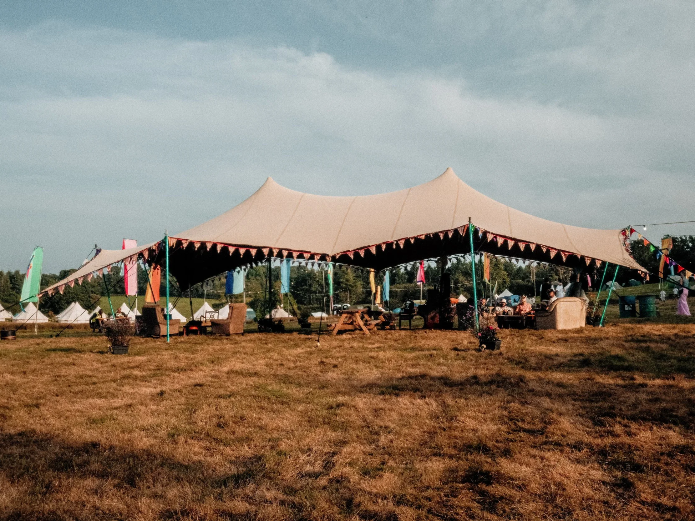 A large outdoor event tent with colorful pennant banners, set on a grassy field under a cloudy sky, with people sitting and standing around.