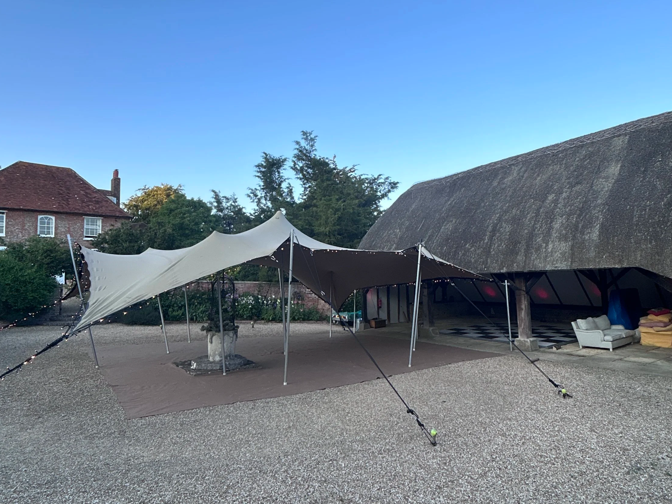 An outdoor scene with a large white canopy tent, string lights, a brick house with a sloped roof, trees, and a barn-like structure, all under a clear blue sky.