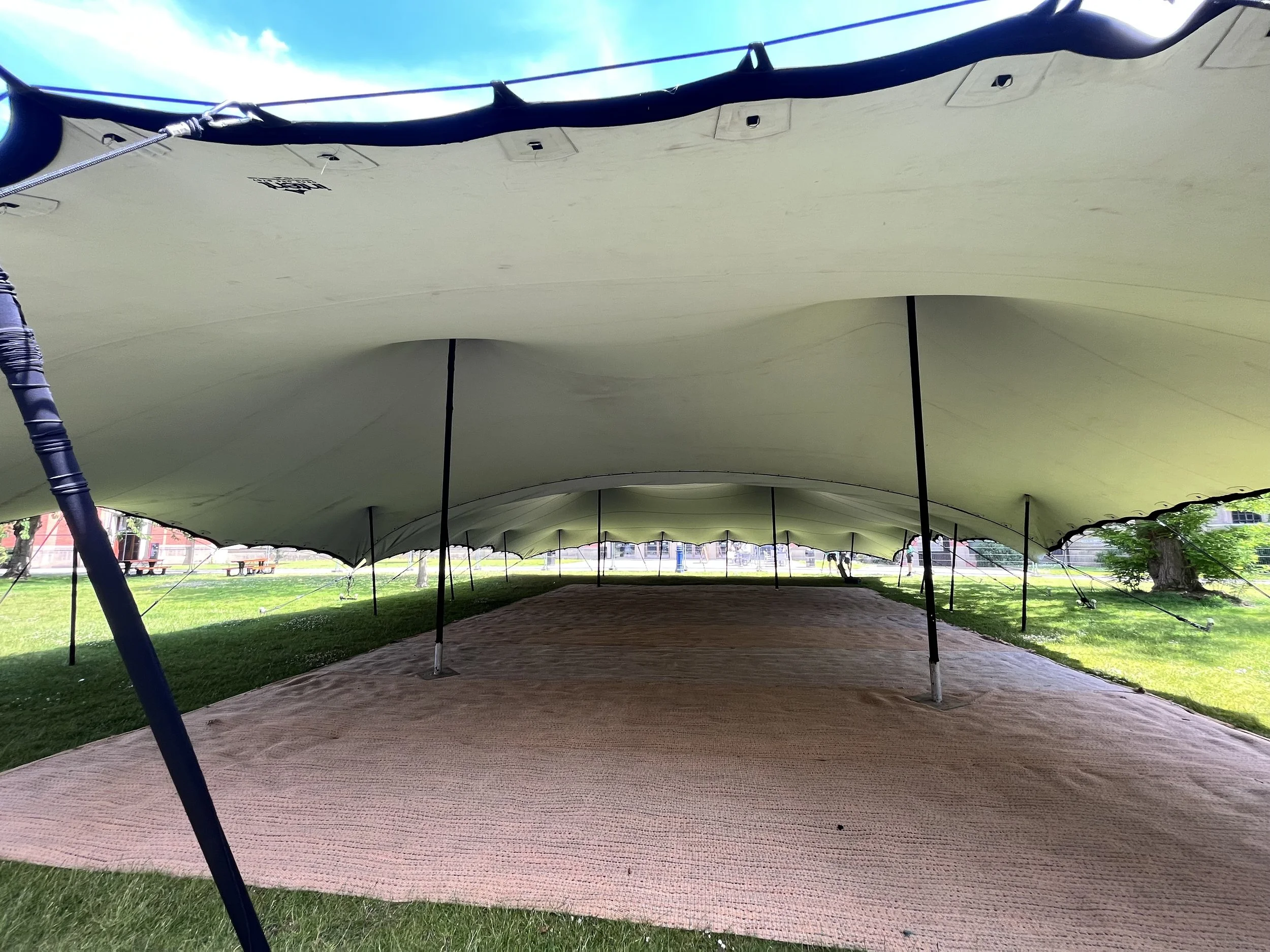 Under a large beige outdoor tent in a park with green grass and trees, there are picnic tables in the background.