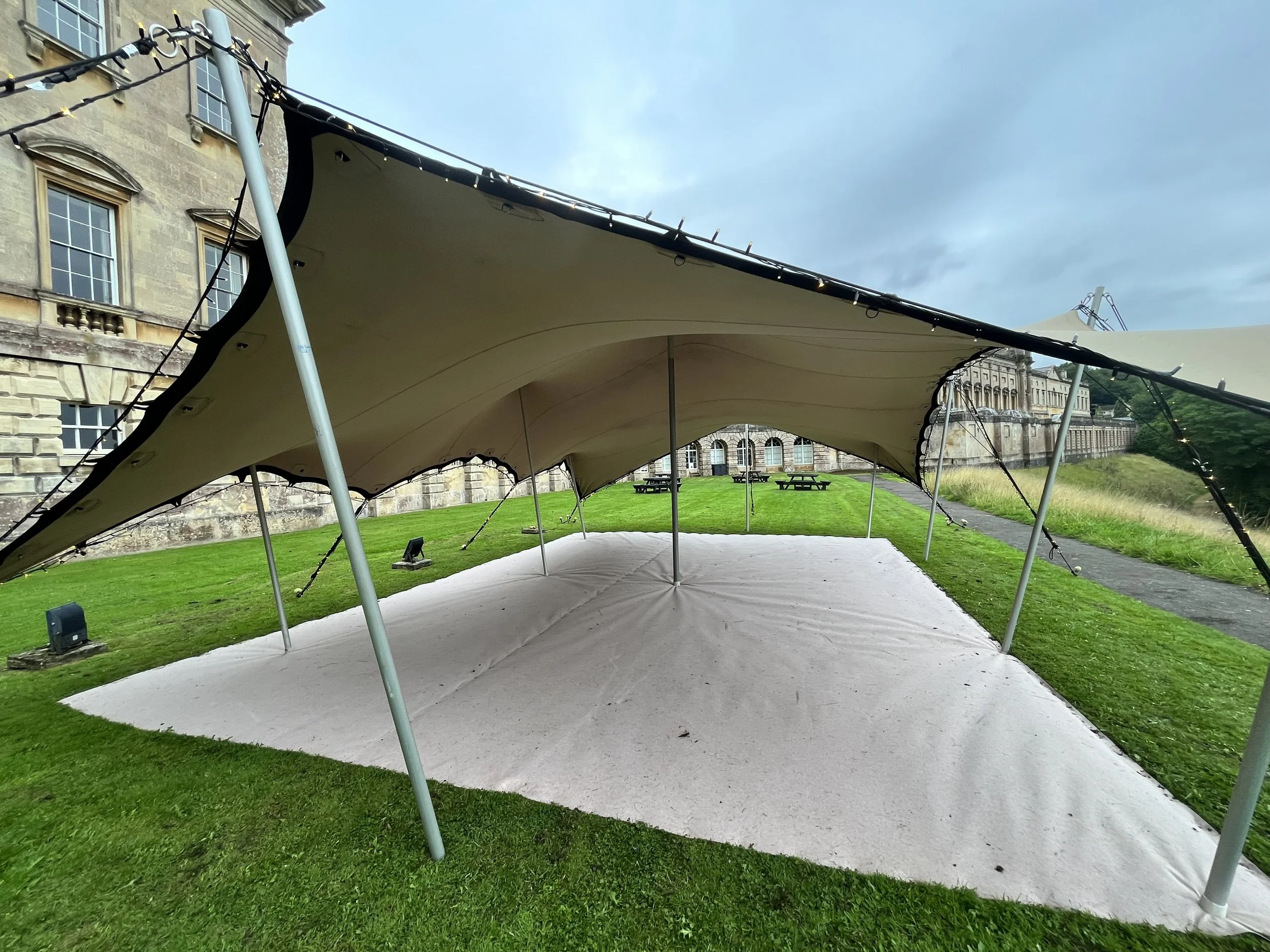 A large outdoor tent or canopy set up on a grassy area in front of a historic stone building, with picnic tables underneath and string lights hanging from the edges of the canopy.