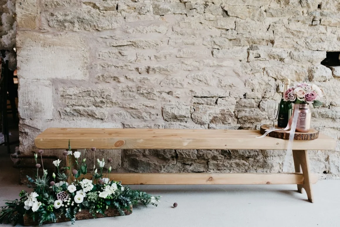 A wooden bench with a flower arrangement and a vase with pink roses on a wood slab, against a textured stone wall.